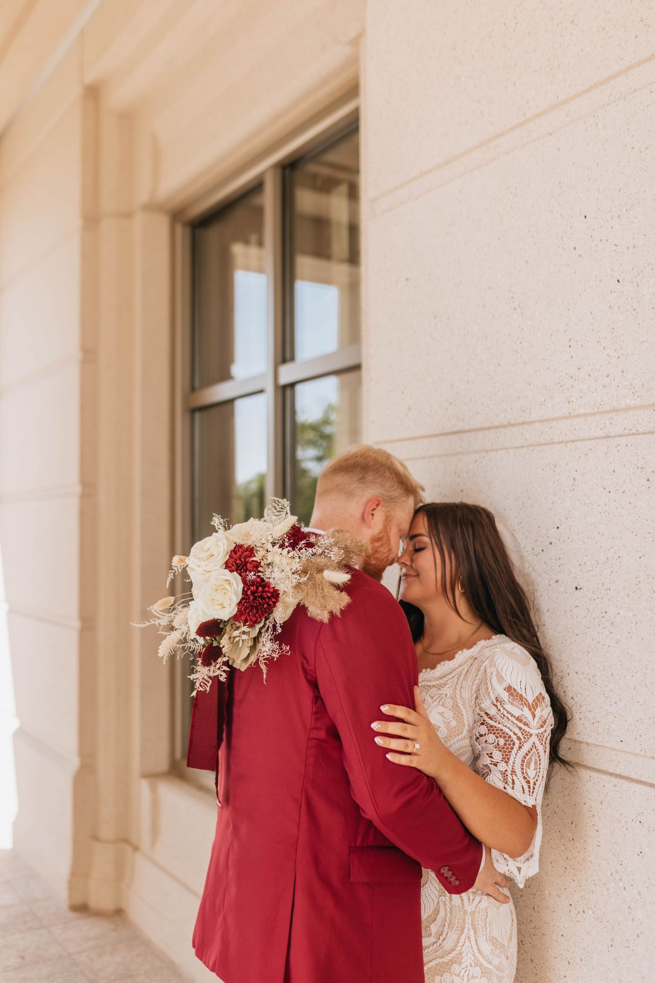 A couple stands close, smiling and touching foreheads, near a window with a stone wall background.