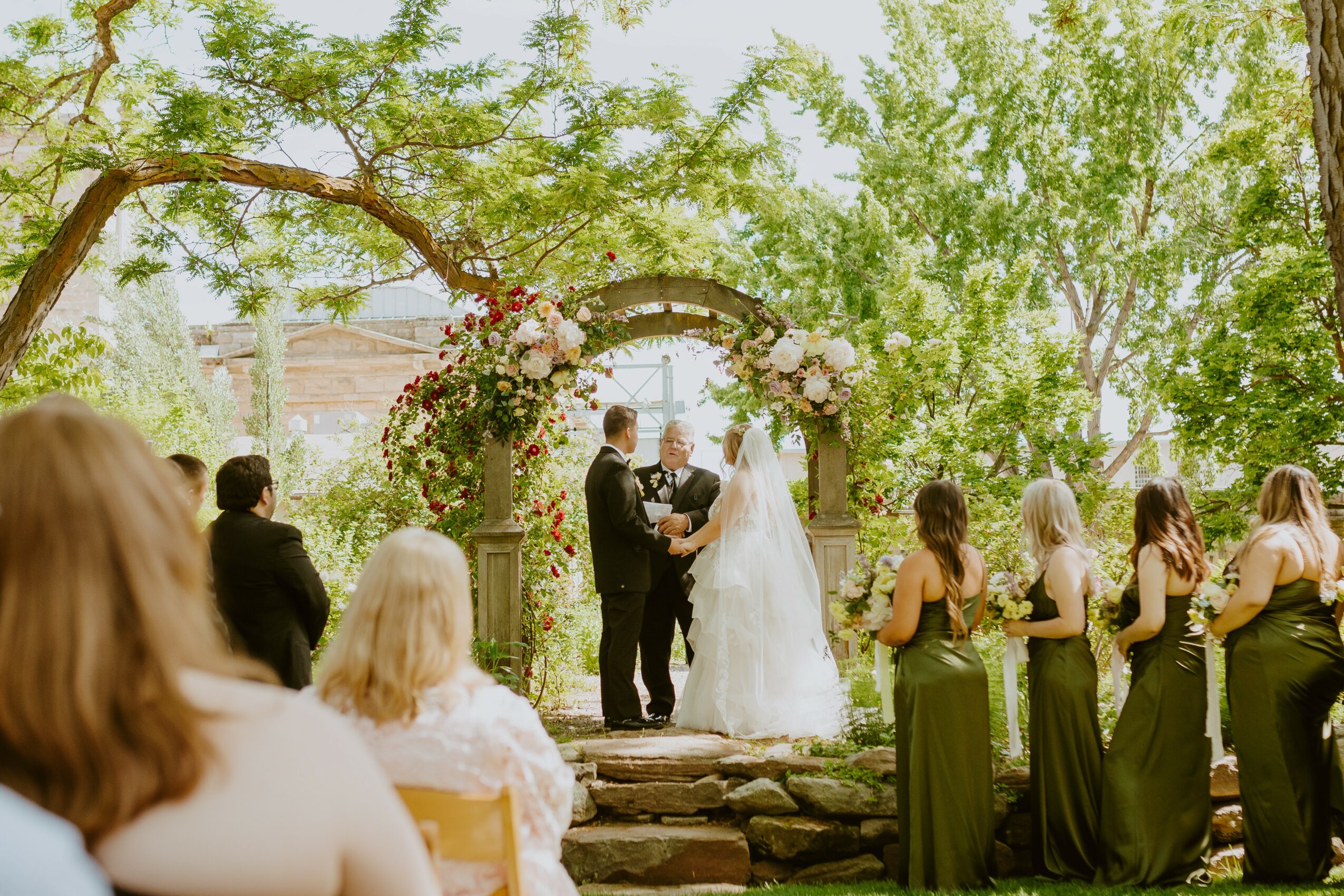 Wedding ceremony outdoors with bride, groom, and guests under a floral arch, surrounded by green trees.