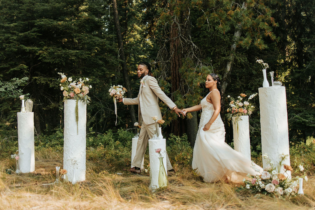 A bride and groom holding hands outdoors surrounded by white pillars with flowers, trees in the background, during a wedding ceremony.