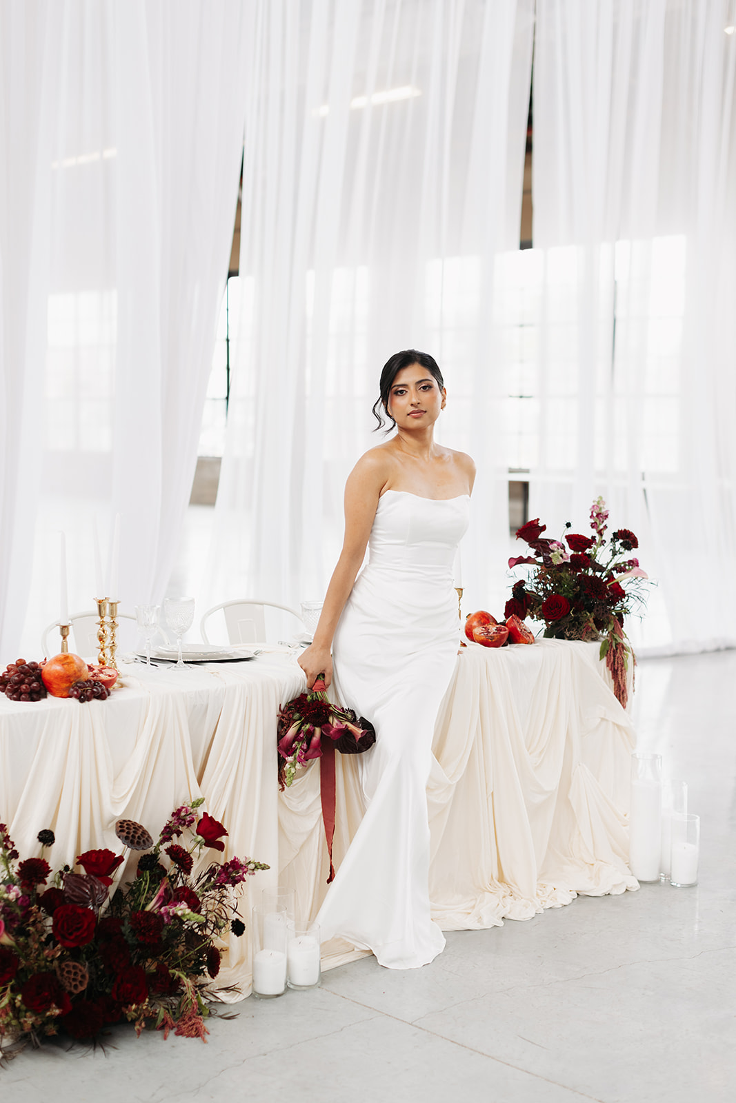 Woman in white dress standing beside tables with flowers and decorations, in a bright room with white curtains
