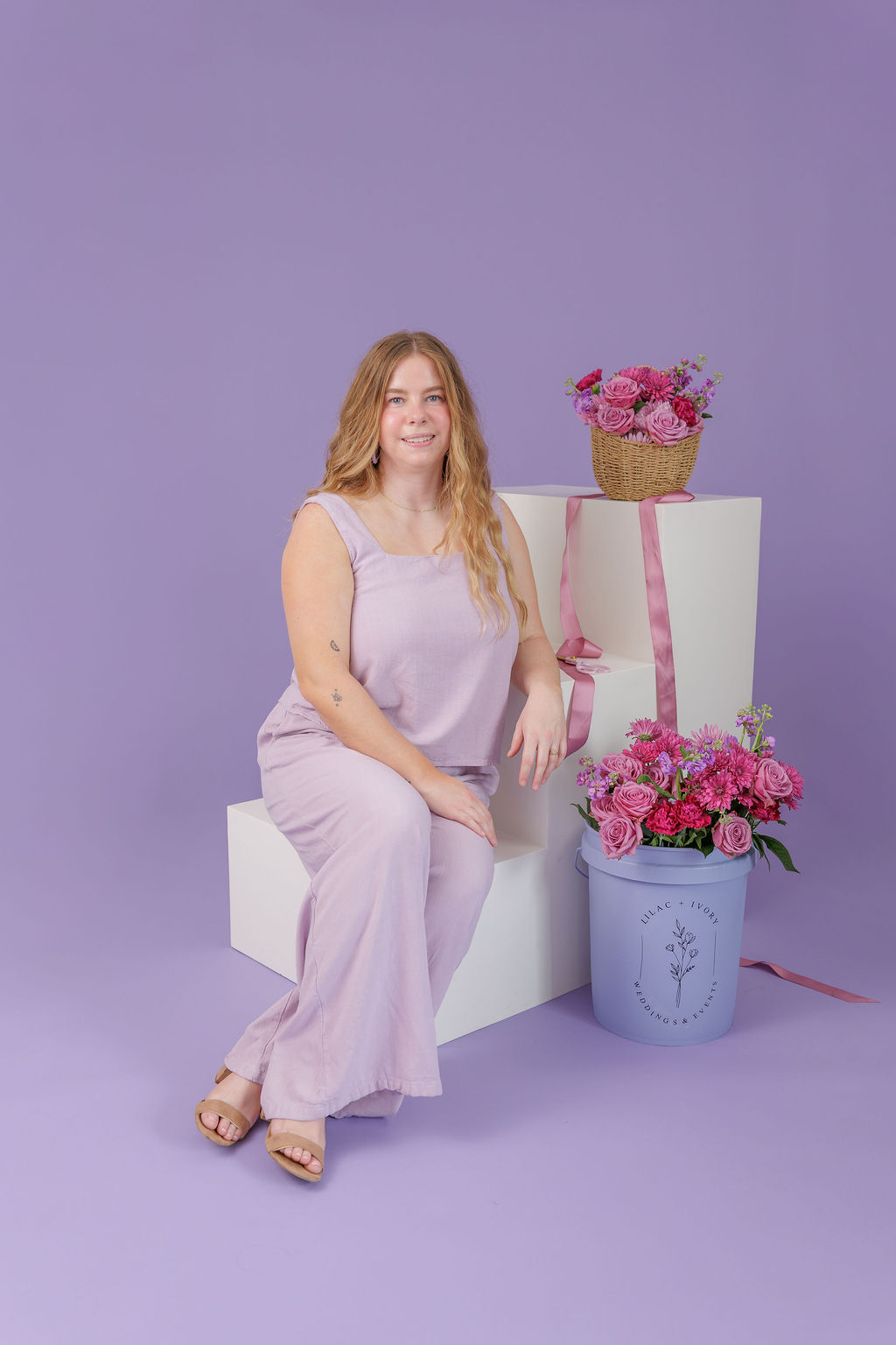 Woman sitting on a white block with pink flowers in baskets and a purple background.