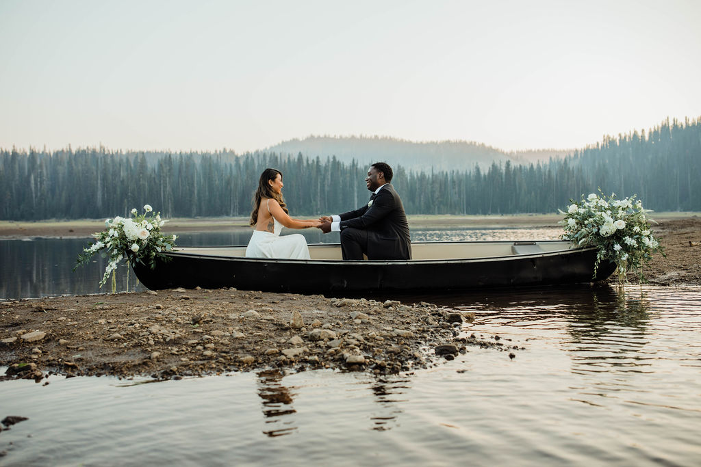 A couple in wedding attire holding hands in a boat on a lake with a forested background.
