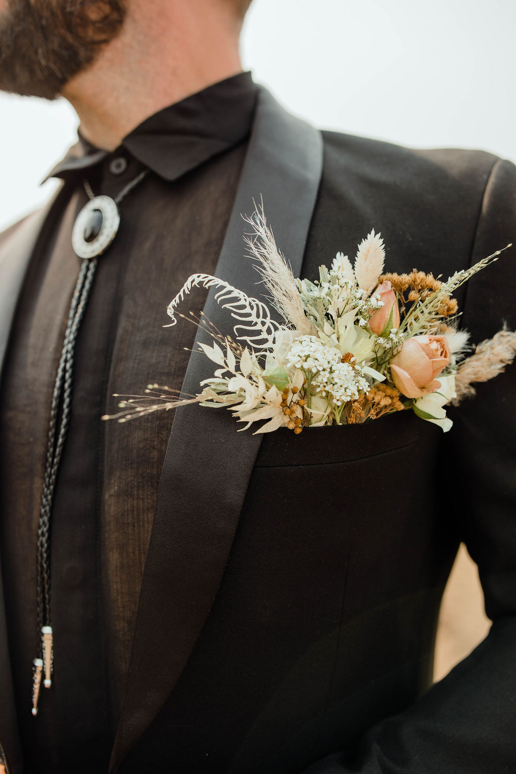 Man in black suit with a floral boutonniere on the lapel, close-up of upper body, neutral background.