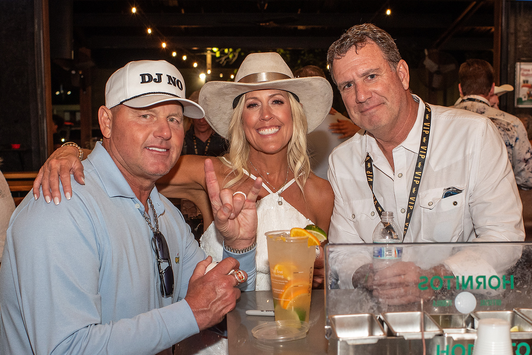 Three people smiling at a table with a drink, one woman wearing a white hat, two men, in a lively setting.