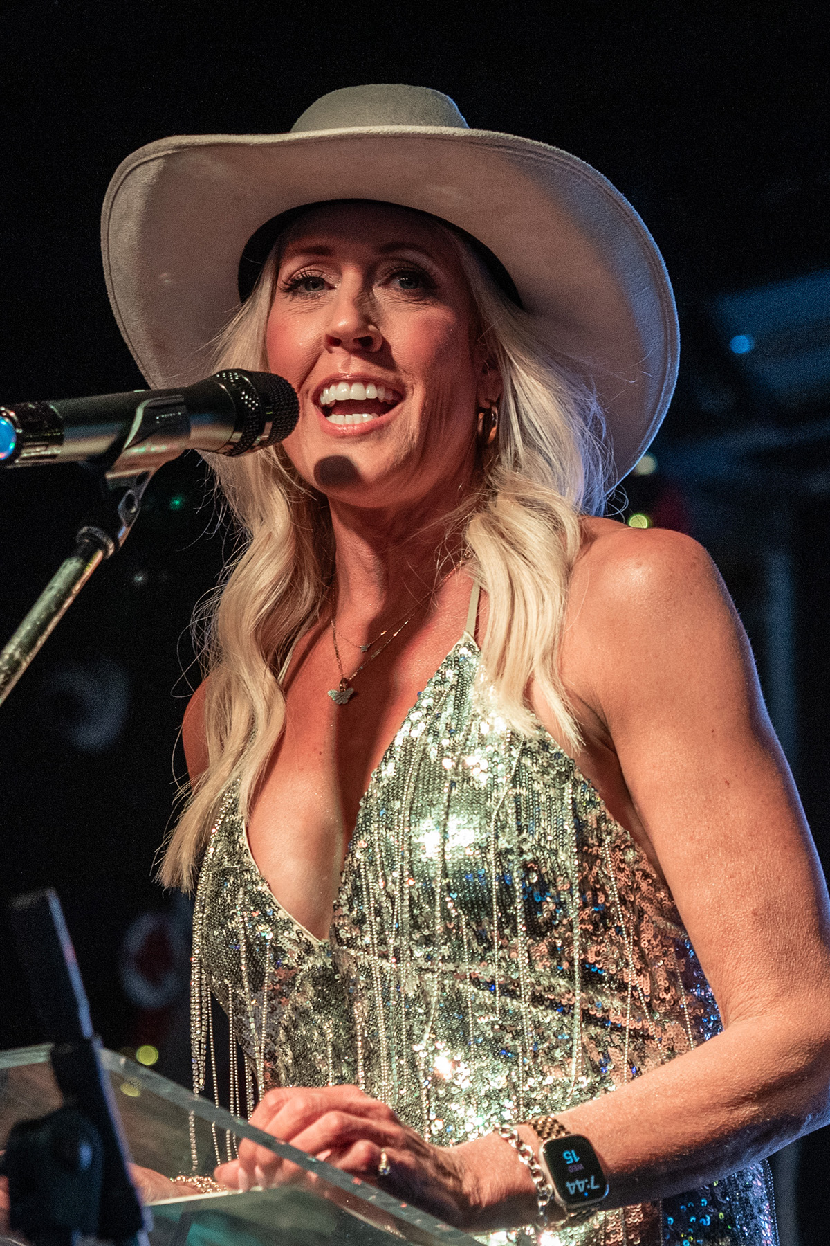 Woman singing into microphone, wearing large hat and sparkly dress, smiling on stage.