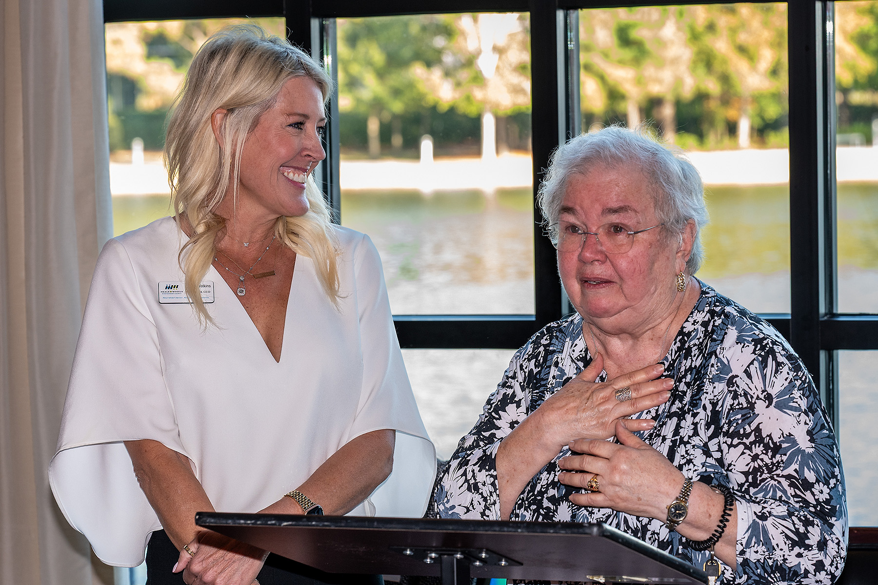A woman in a white uniform smiling and talking to an elderly woman with glasses, sitting at a table near a window with a view of water and trees.
