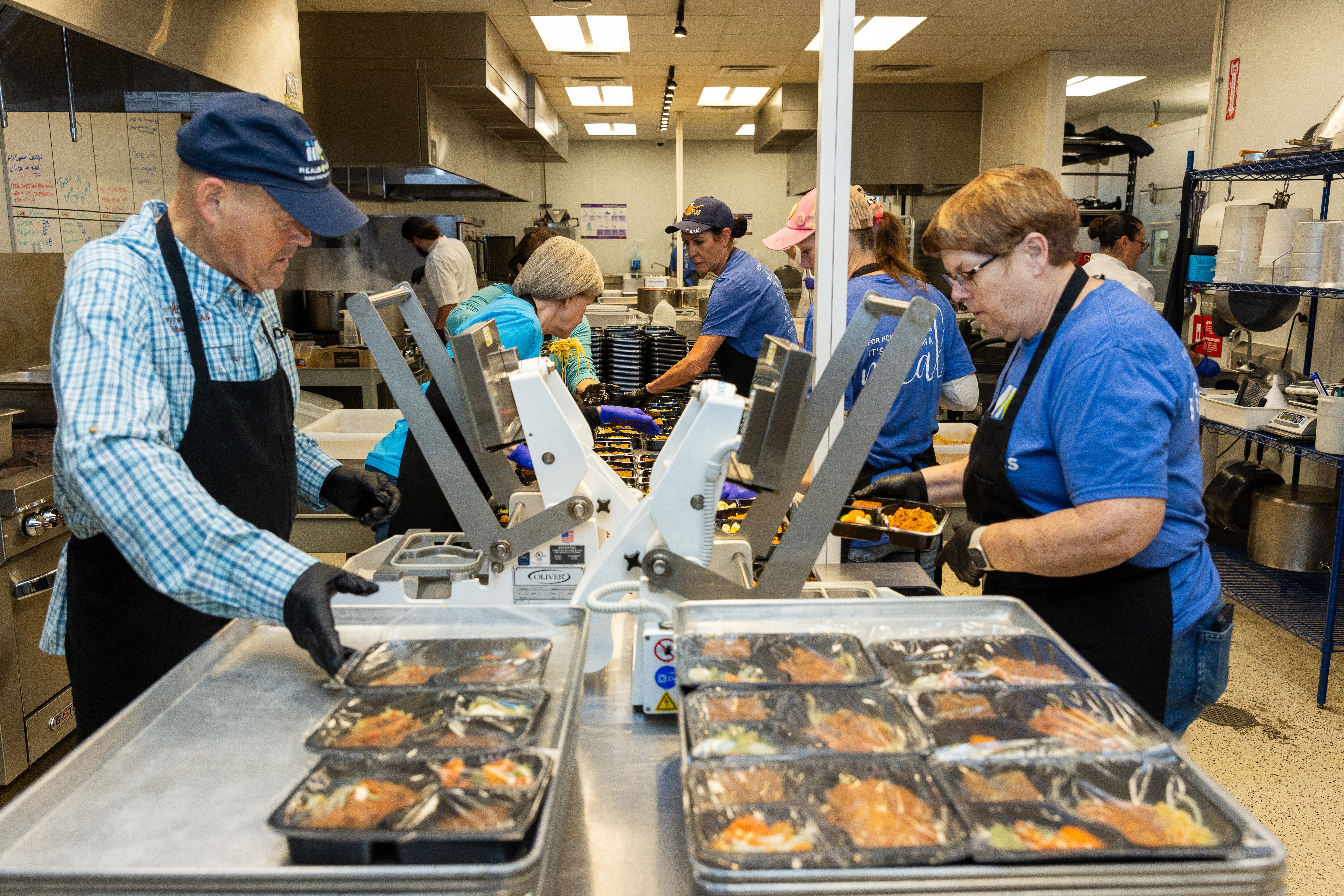 People working in a food packaging facility with trays of prepared meals on a conveyor belt.