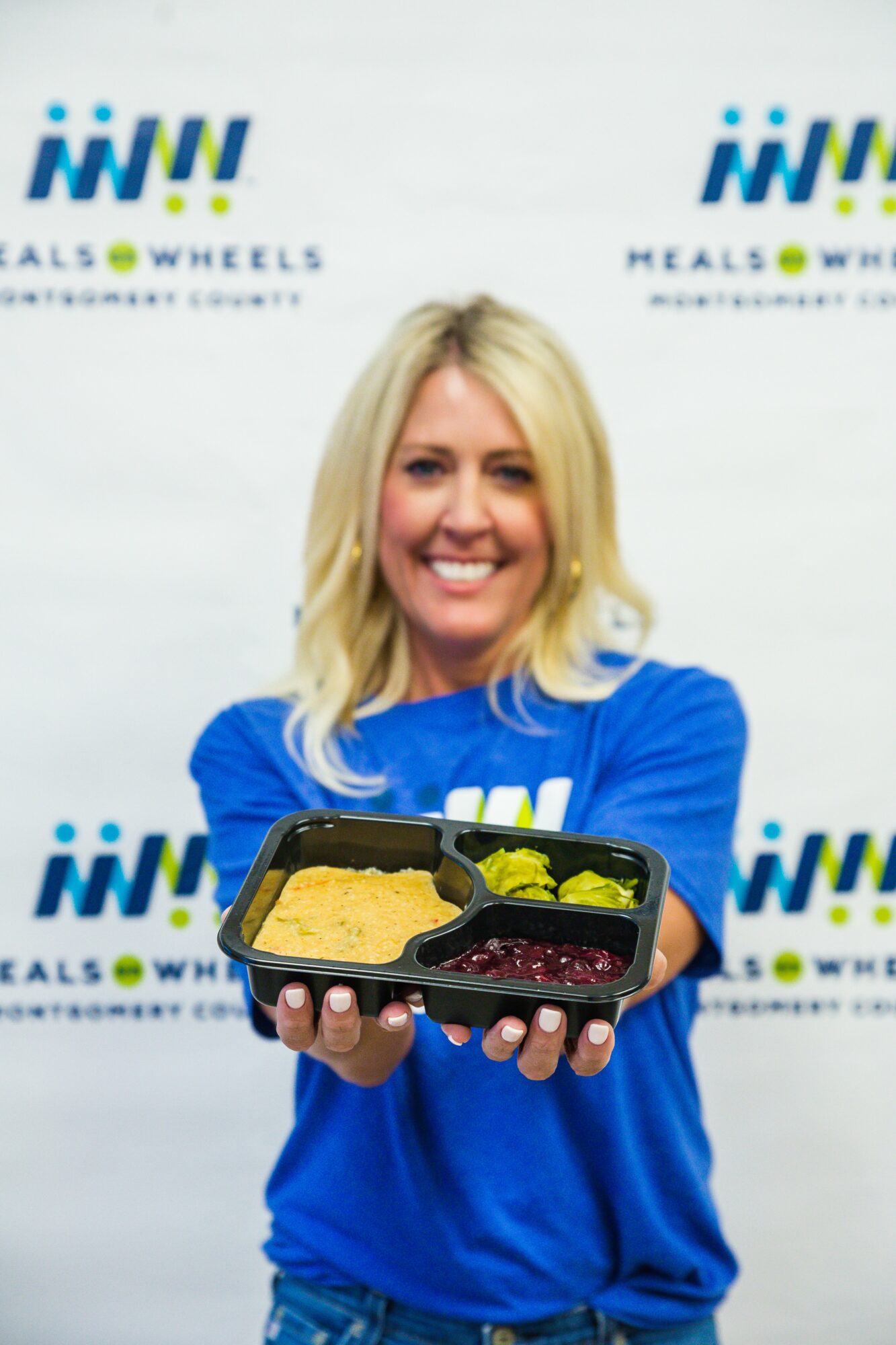 Woman with blonde hair in a blue shirt holding a meal tray with food, smiling, background with logos.