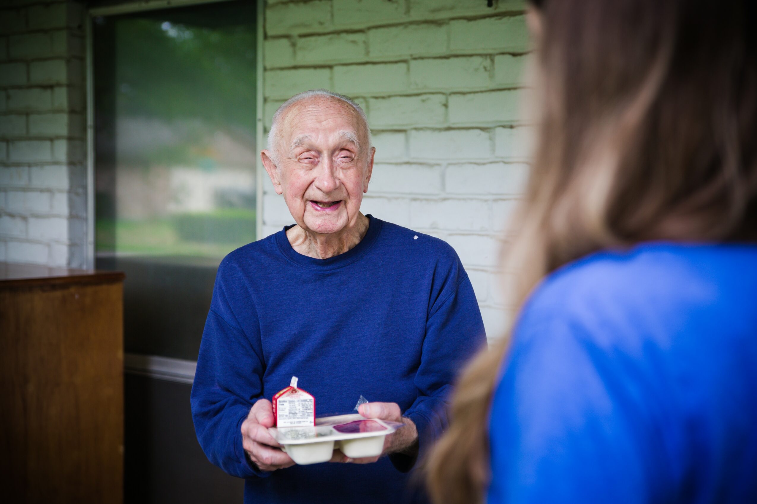 Elderly person smiling and holding a tray with a small cake, talking to a person with long hair, outside a building