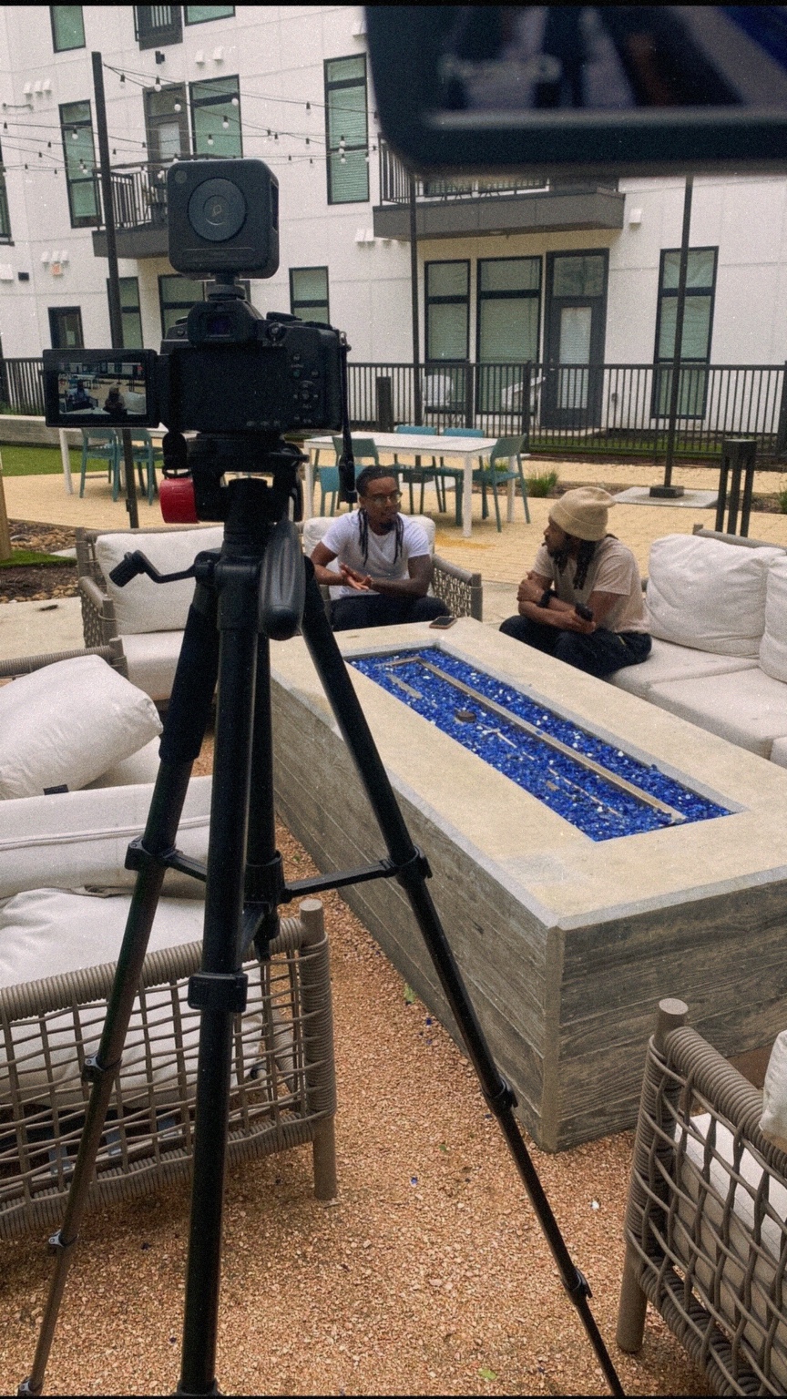 Two people sit at a table outdoors with a water feature, while a camera on a tripod is in the foreground.