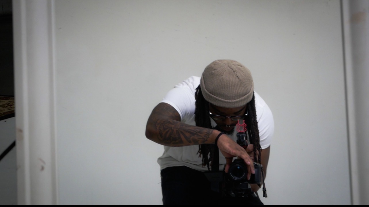 Person with a beige head covering and tattoos, holding a camera, leaning forward against a plain white background.