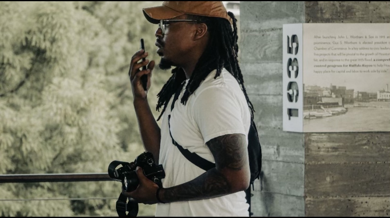 Person with glasses, cap, and backpack talking on a phone outdoors, holding a camera, with trees in the background.