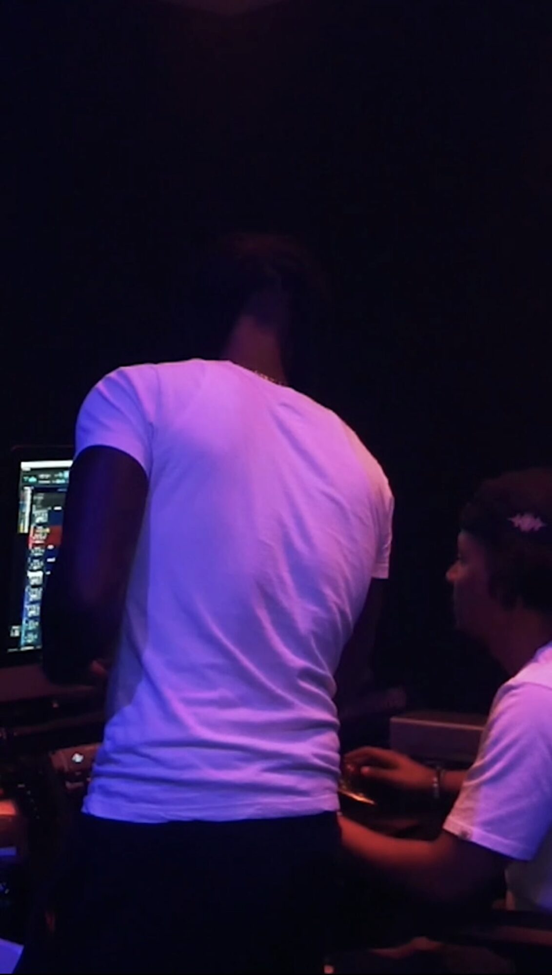 Two people in a dark room with electronic equipment, one standing and one sitting, both wearing white shirts.