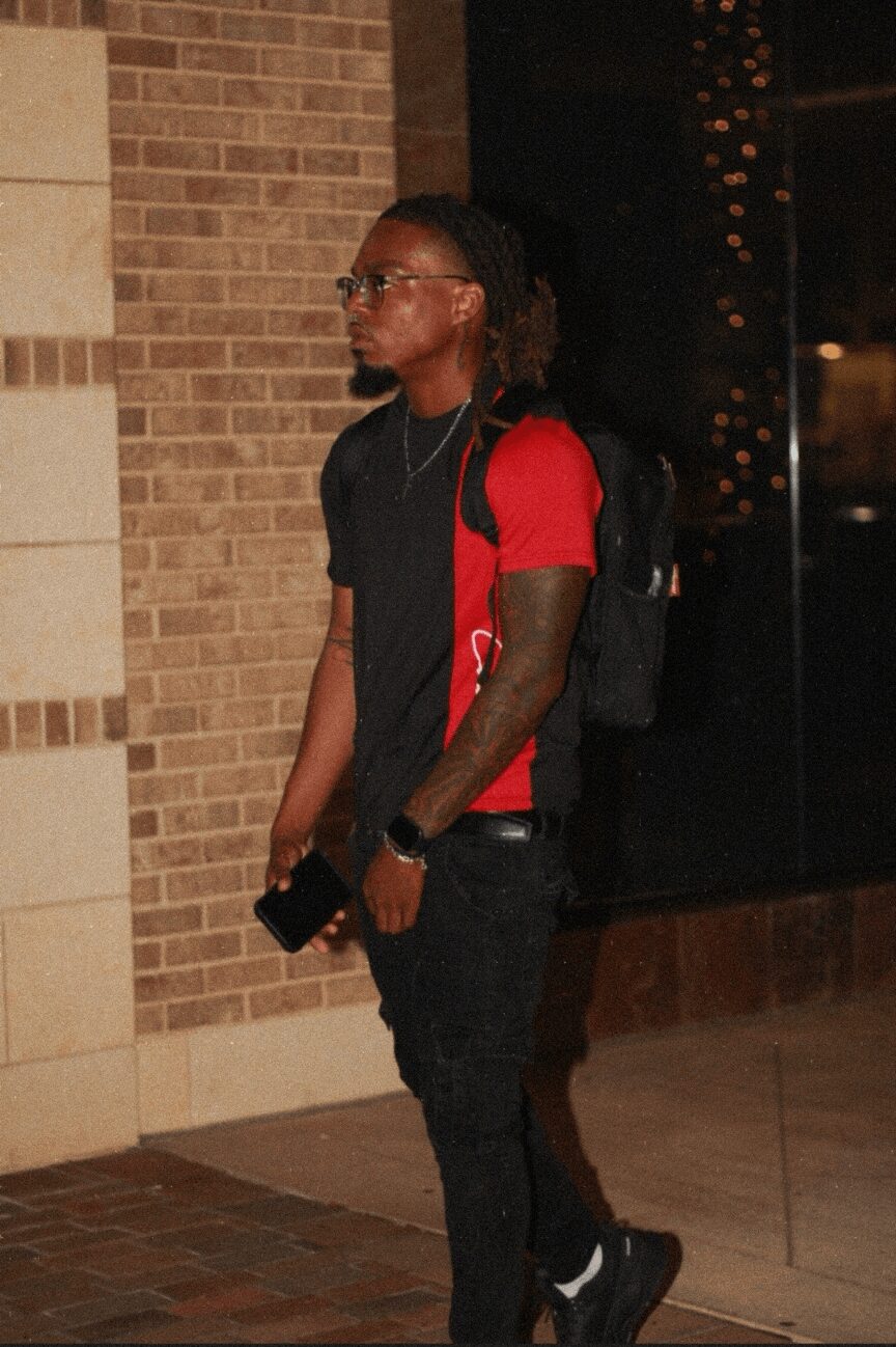 Young man with glasses, dreadlocks, red and black shirt, backpack, standing against brick wall at night.