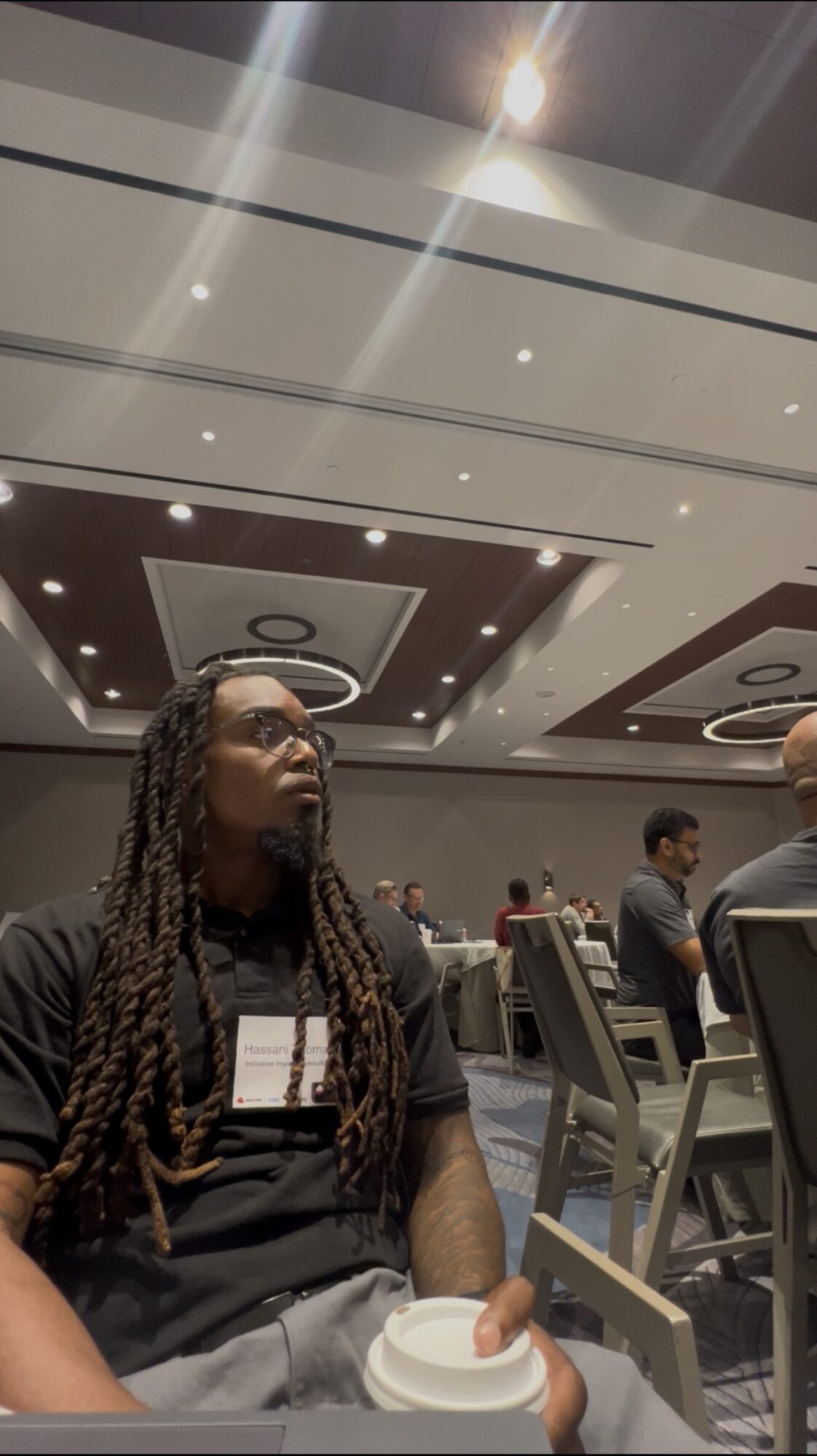 Person with long dreadlocks wearing glasses and a black shirt, sitting at a table in a conference room.