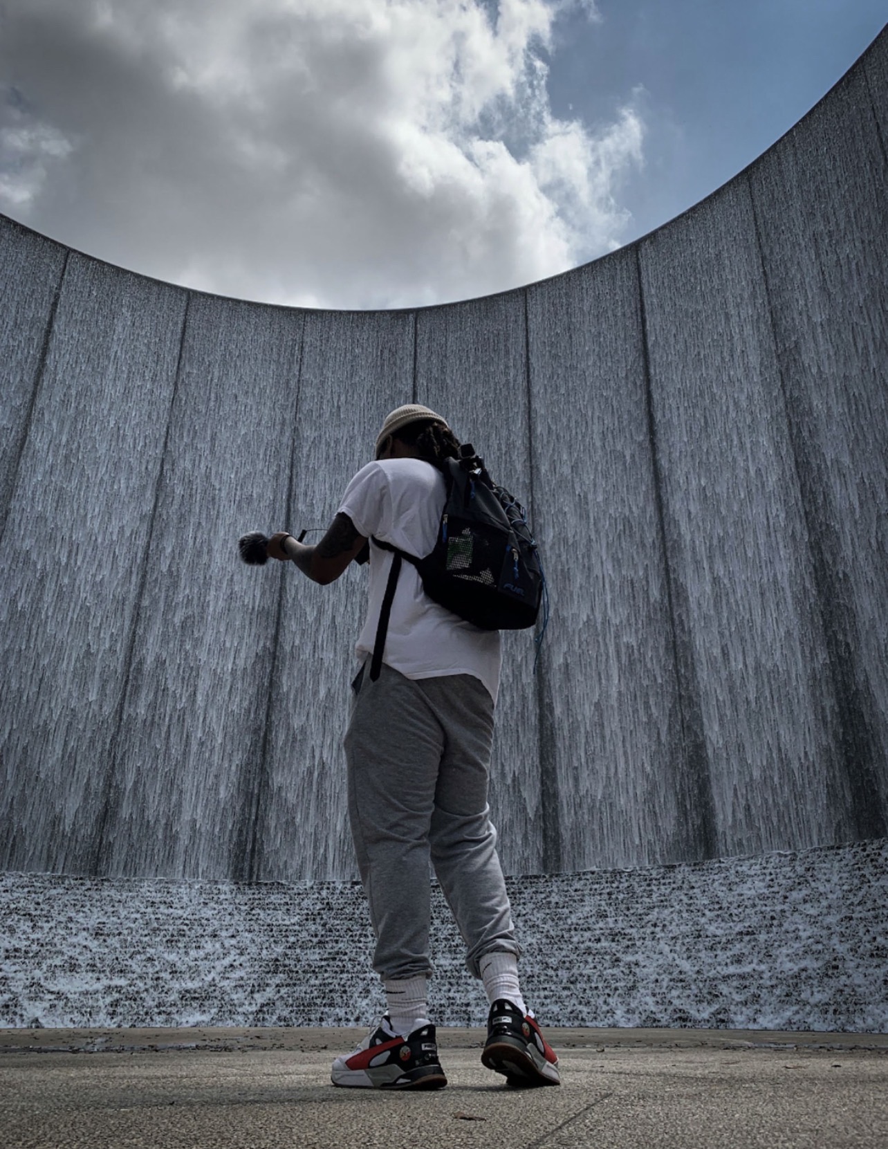 Person with backpack and helmet standing in front of a curved concrete wall with water flowing down, sky with clouds above.