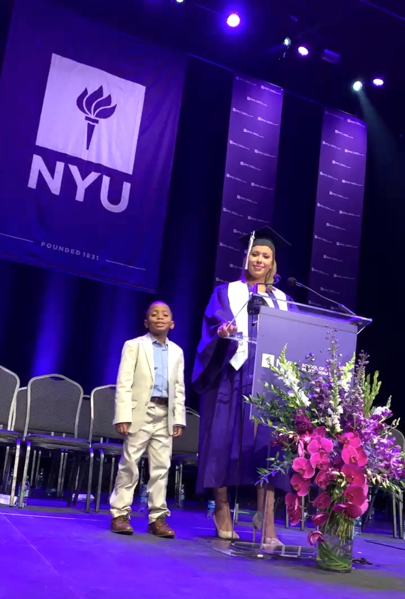 Woman in purple graduation gown speaking at podium with a young boy in a suit on stage, NYU banner in background, flowers in front.