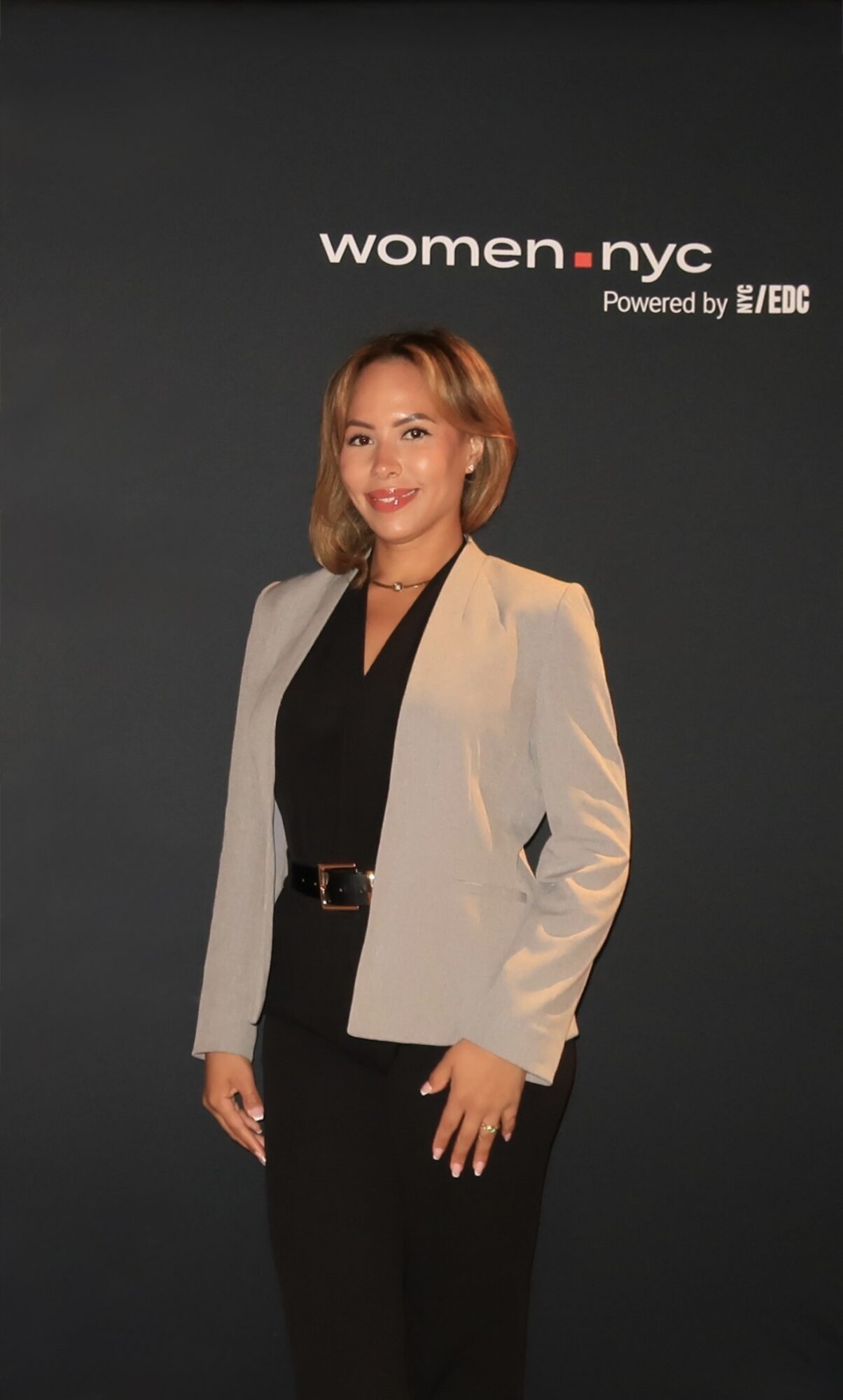 Woman with shoulder-length hair in a beige blazer and black outfit, standing in front of a dark background with 'women.nyc' text.