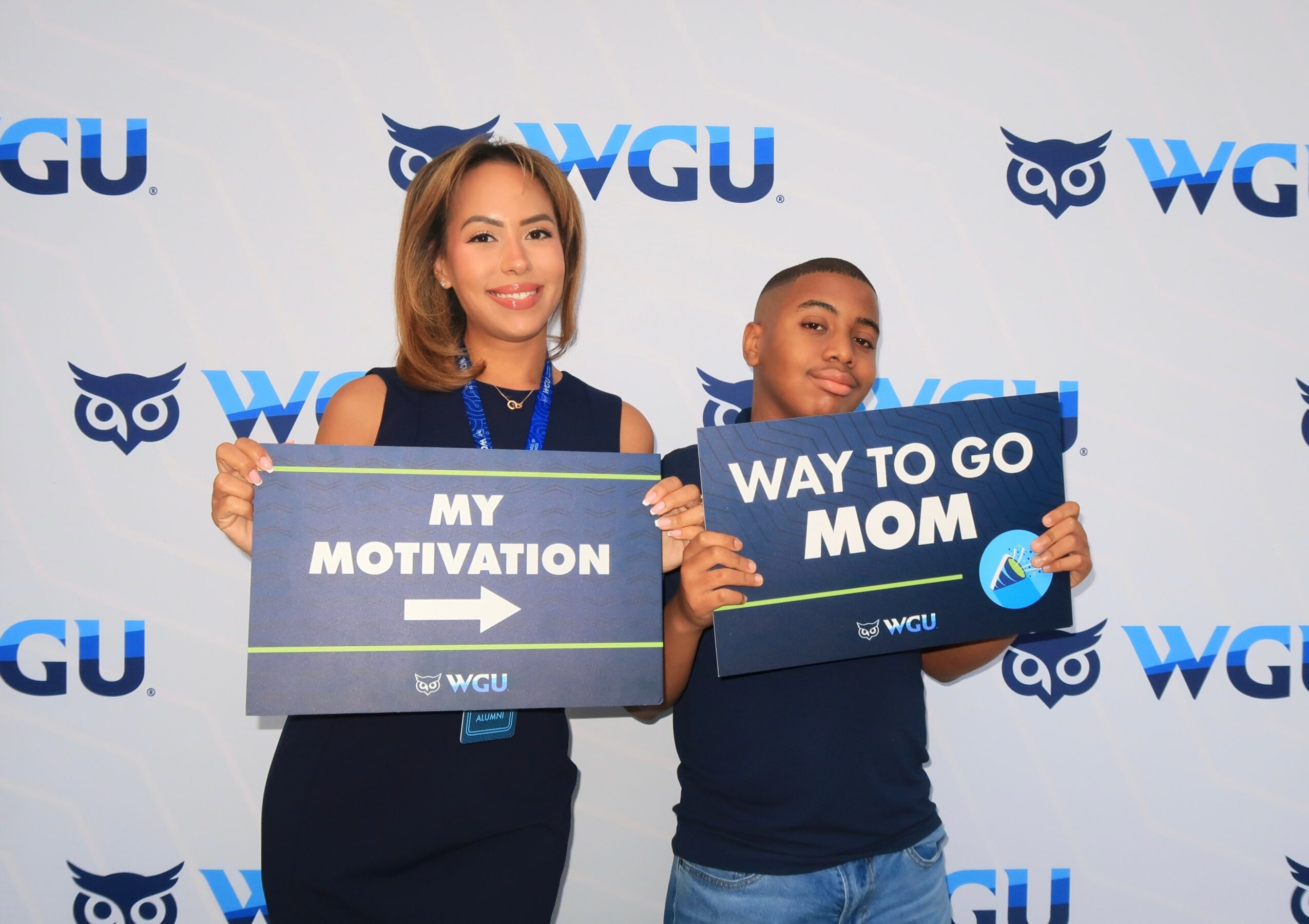 Two women holding signs at an event with WGU logos in the background, smiling. One sign says 'My Motivation,' the other 'Way to go Mom.'