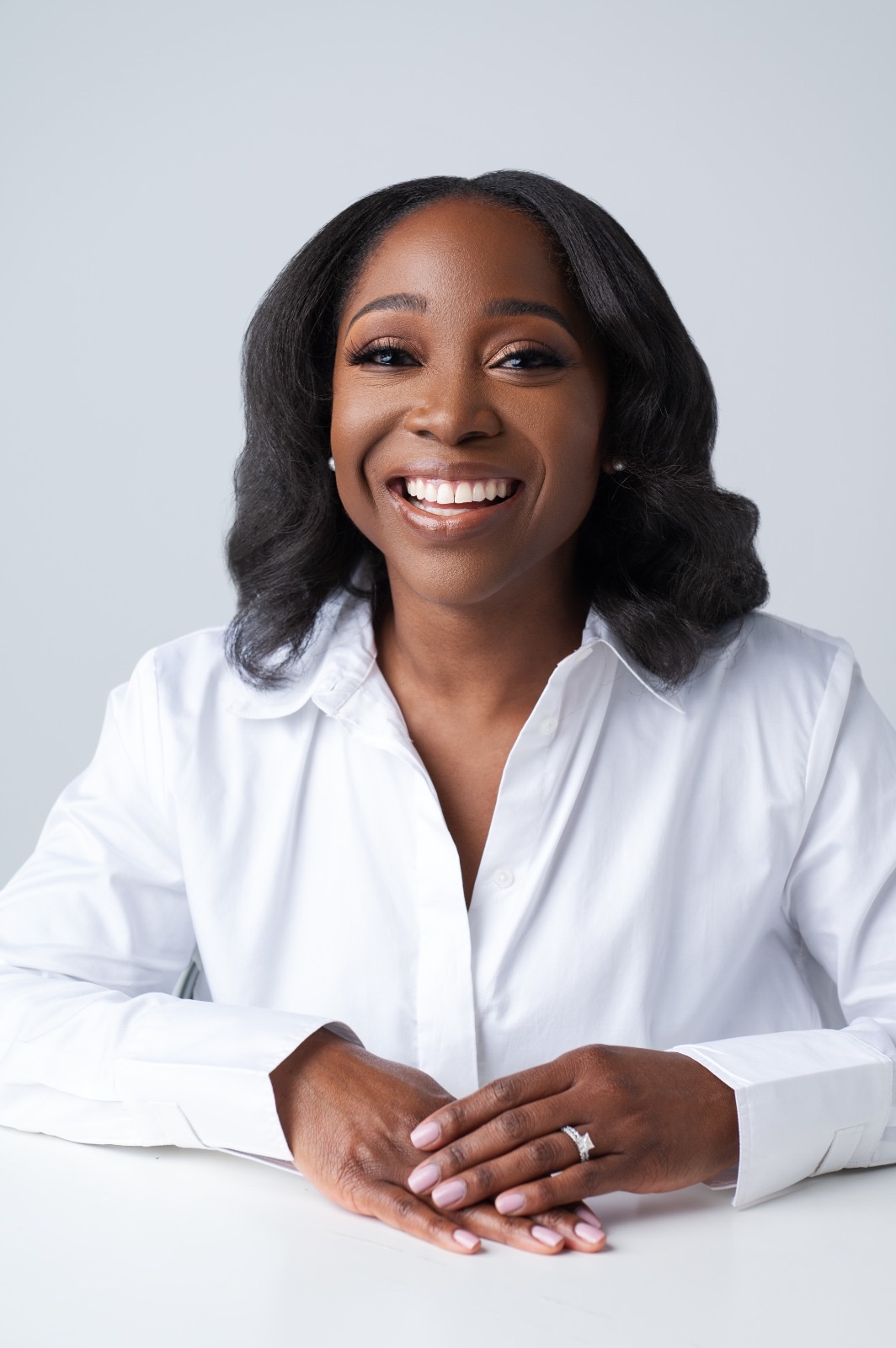 Smiling woman with shoulder-length wavy hair wearing a white shirt, sitting with hands folded on a table, neutral background.
