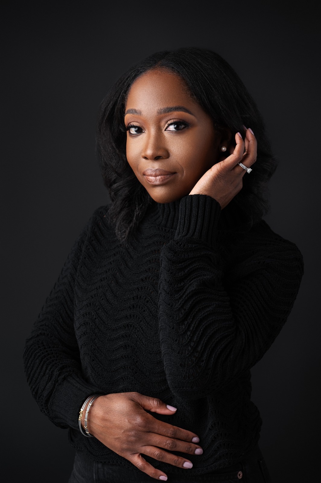 Portrait of a woman with dark hair wearing a black sweater, posing with her hand near her face against a dark background.