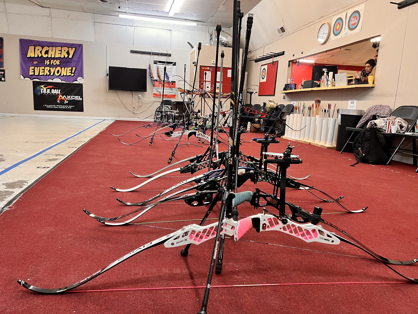 Indoor archery range with bows lined up on a red carpet, targets on the wall, and a sign that says 