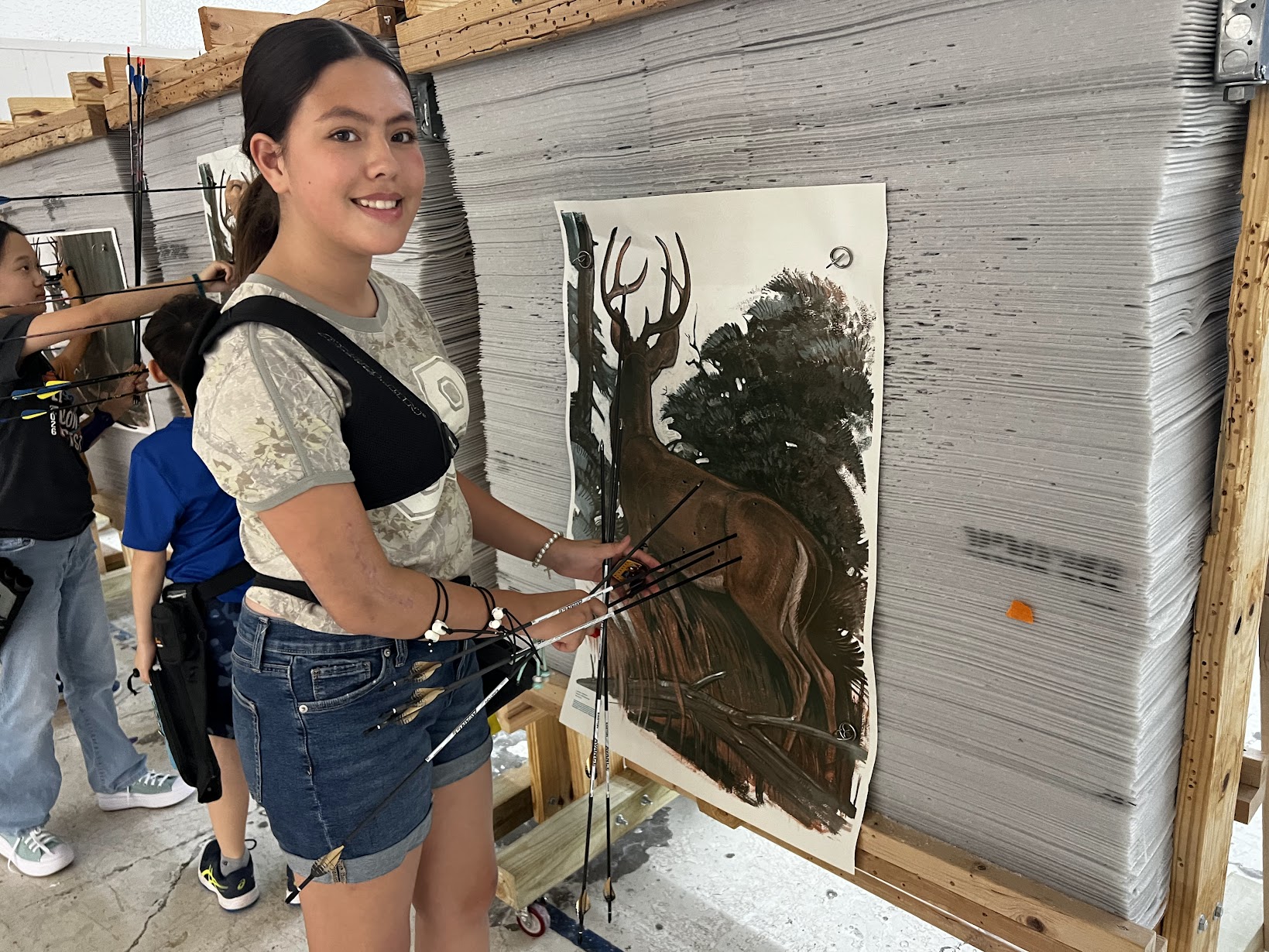 Young woman smiling, holding paintbrushes, standing next to a painting of a deer in a forest, in an art studio.