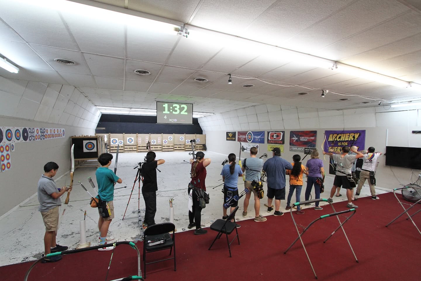 People line up at an indoor shooting range, aiming at targets on the wall, with a digital sign displaying '132'.