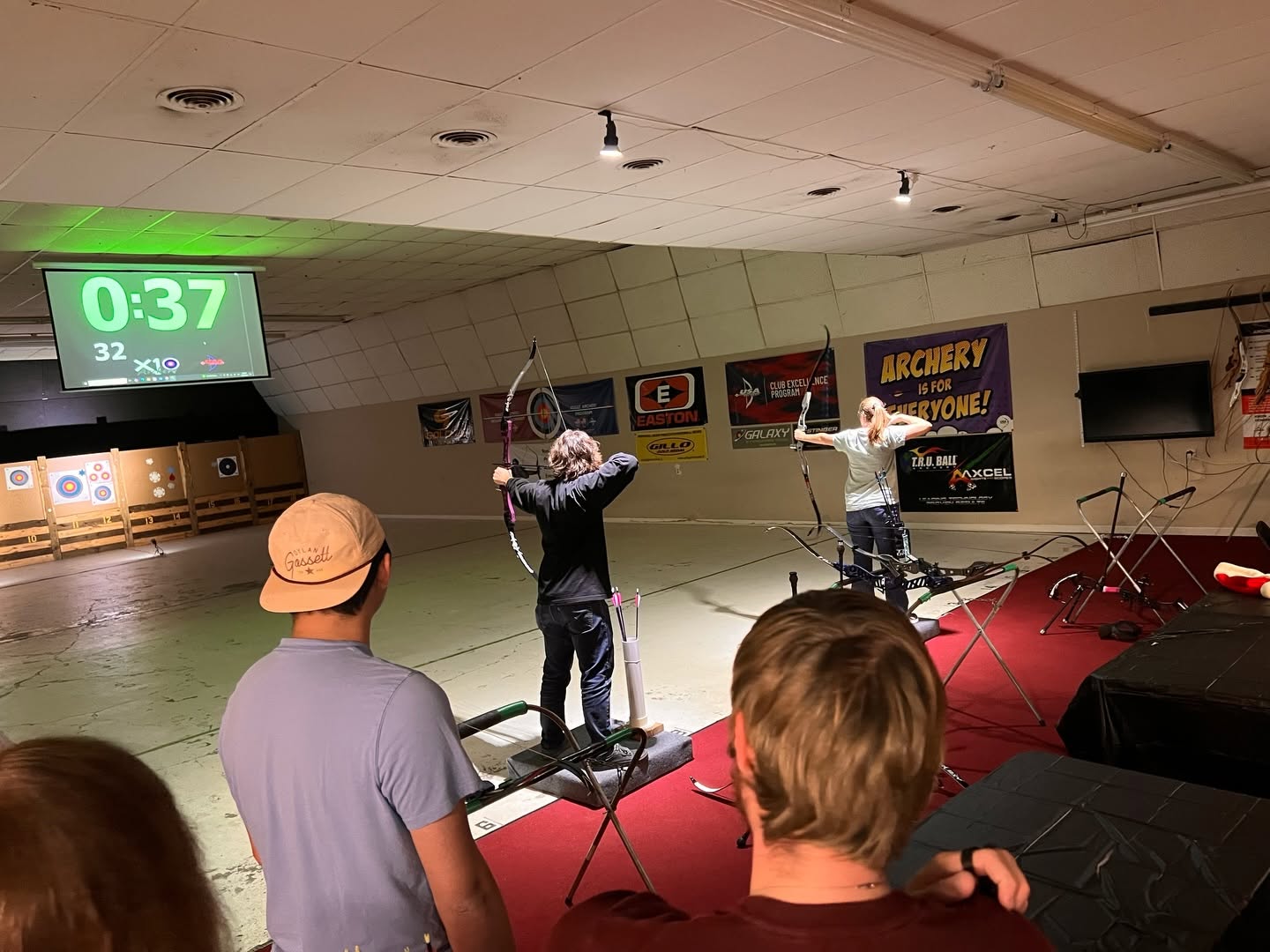 Indoor archery range with two people shooting bows, others watching, digital timer showing 0:37, and various posters on the wall.