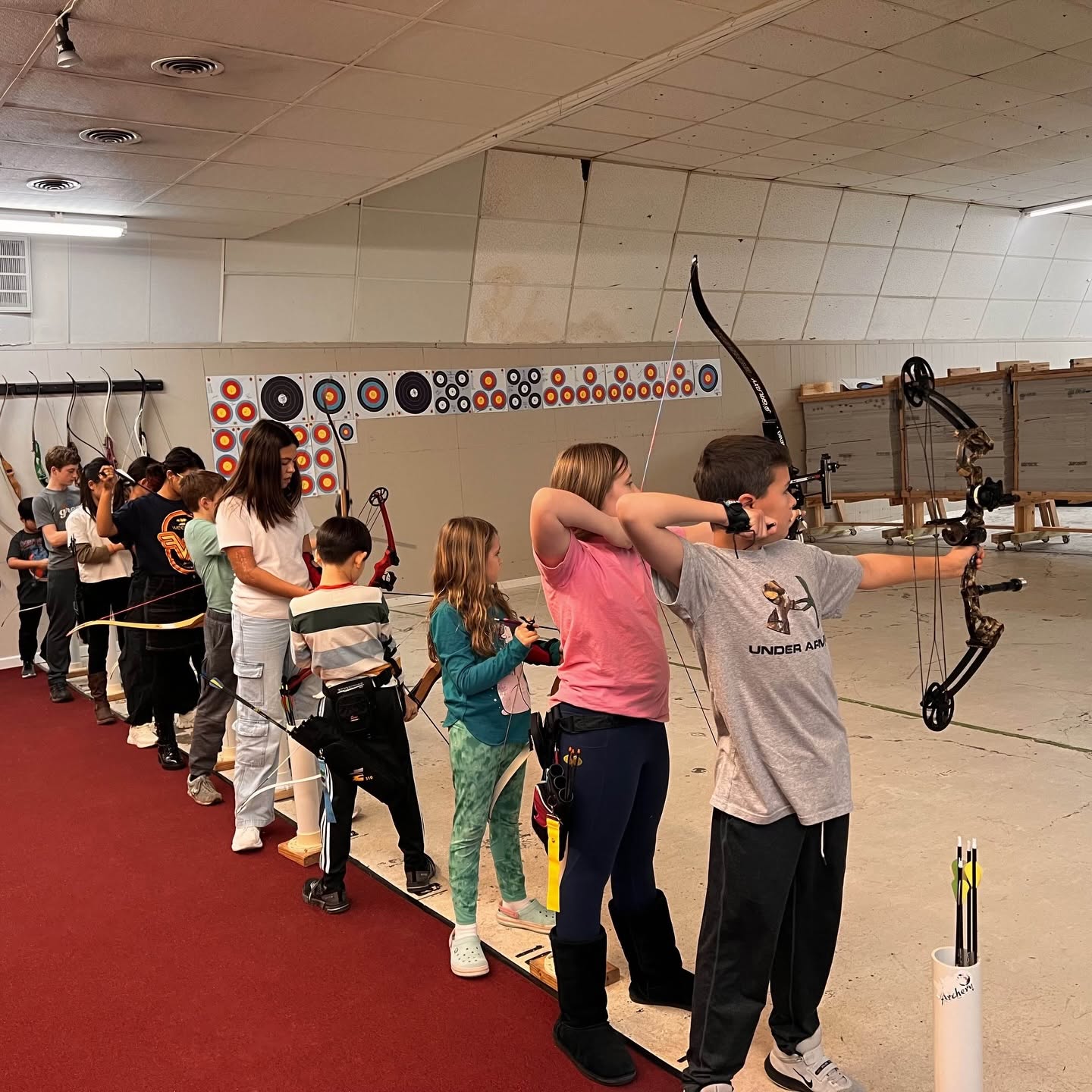 Group of children and a woman practicing archery indoors, with targets on the wall and a red carpeted floor.