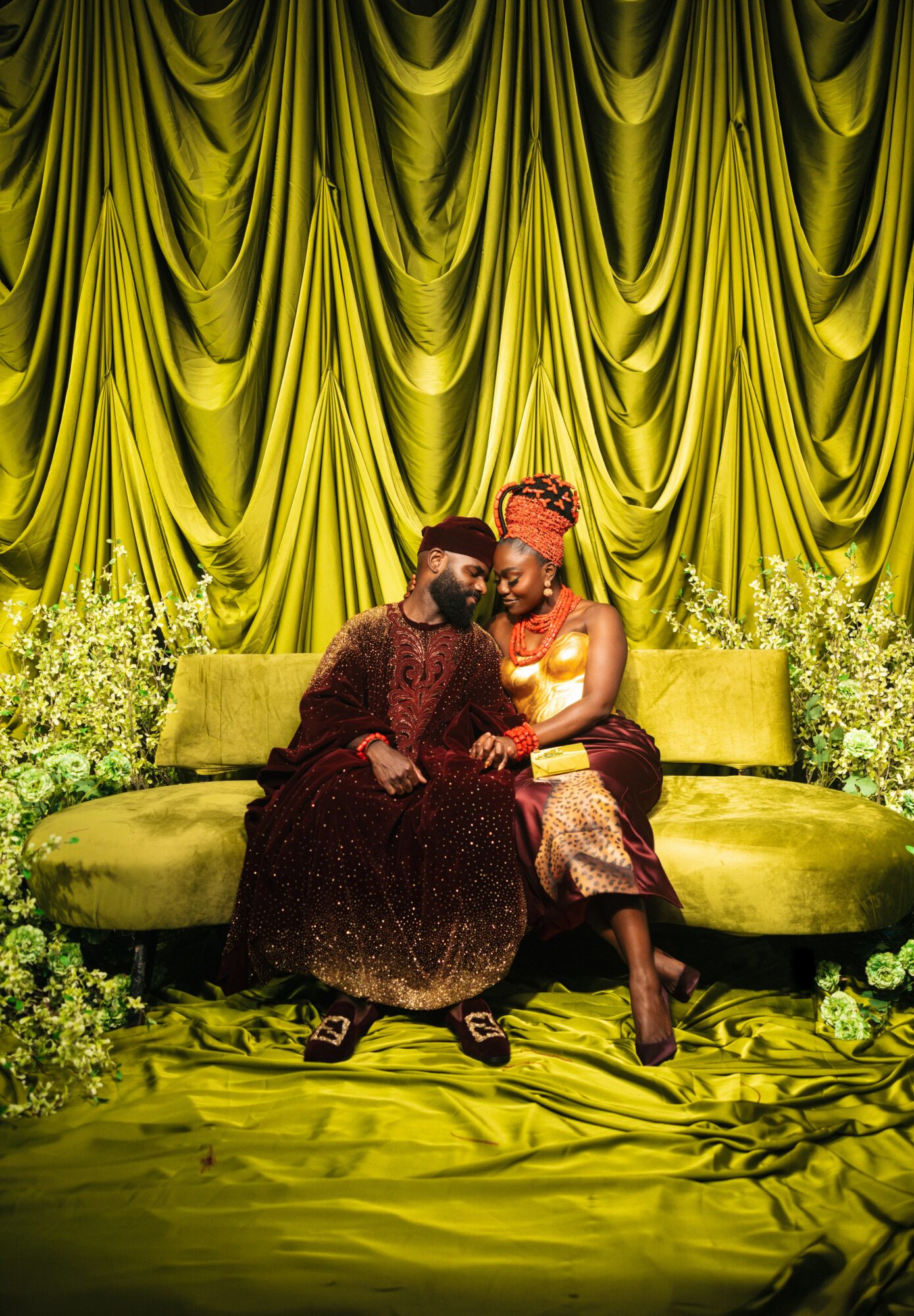 Two people sit on a green velvet sofa with a yellow curtain backdrop, surrounded by flowers, in formal attire.