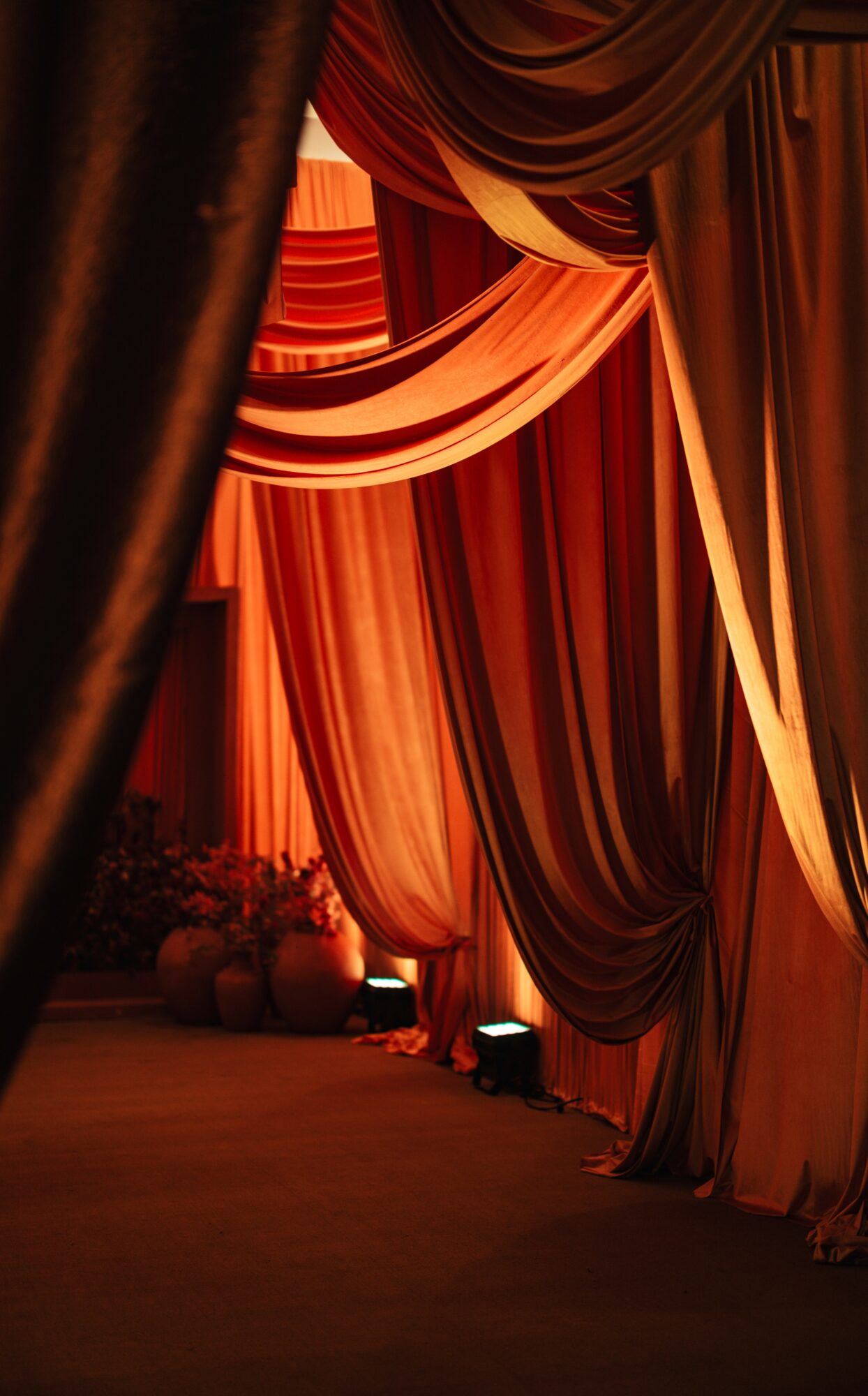 Curtains and drapes in warm colors hang in a decorated setting, with soft lighting and floral arrangements in the background.