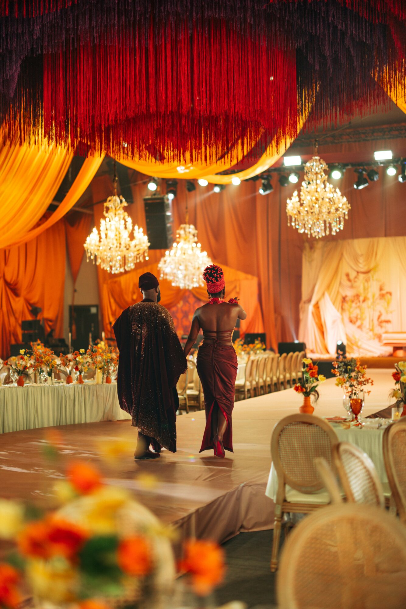 Two women walk on a decorated stage with chandeliers and draped fabric, in a banquet hall setting.