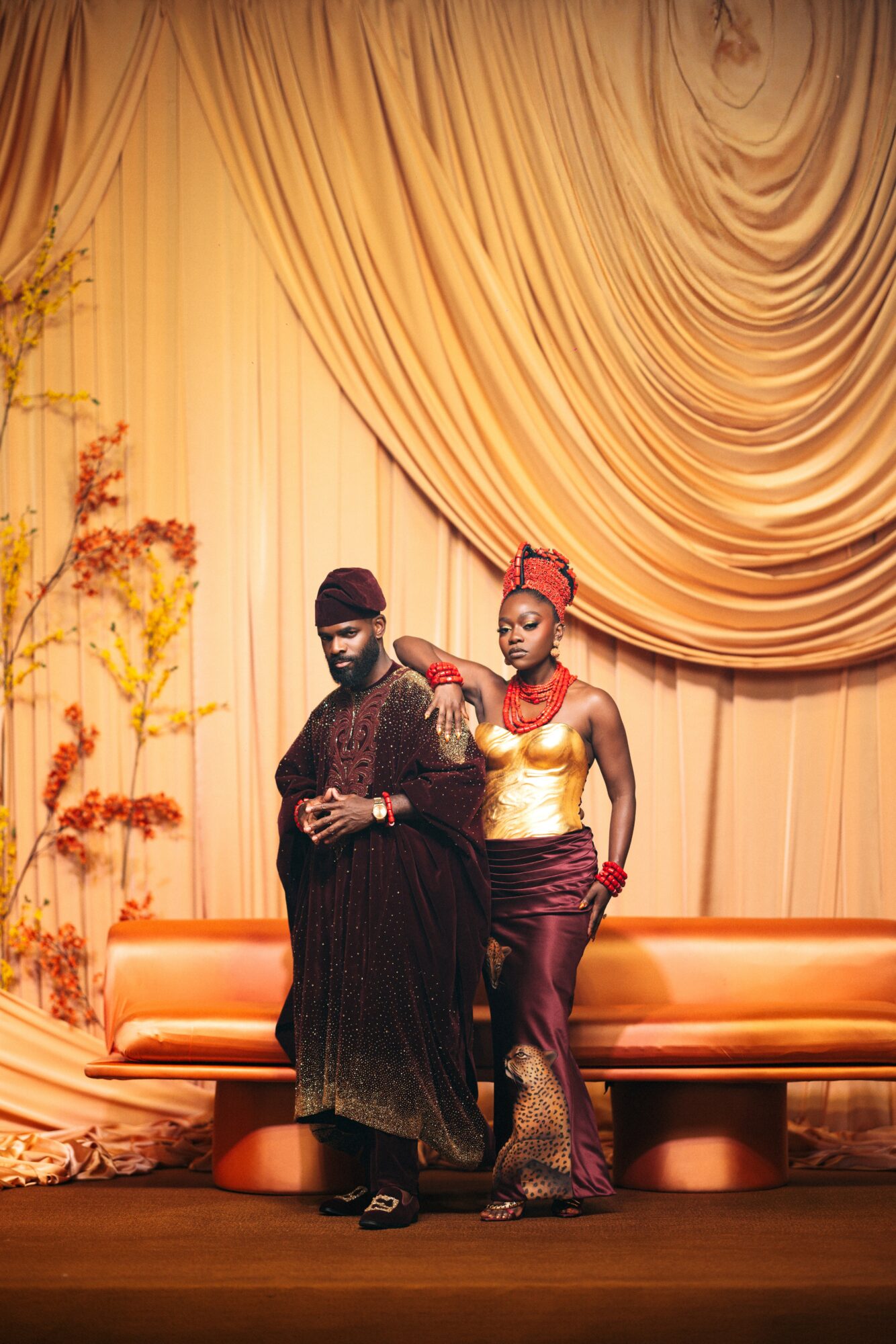 Two people stand on stage in front of a gold curtain, one in traditional attire and the other in a gold top and maroon skirt.