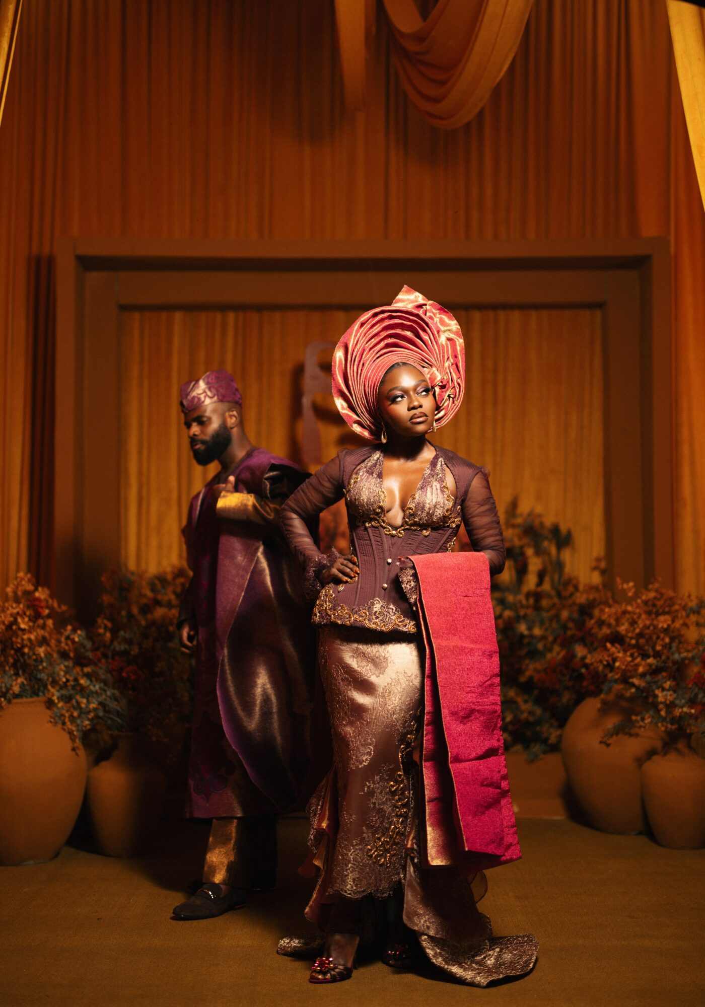 Two women in traditional attire on stage with floral arrangements, one holding a pink cloth, in front of a curtain backdrop.