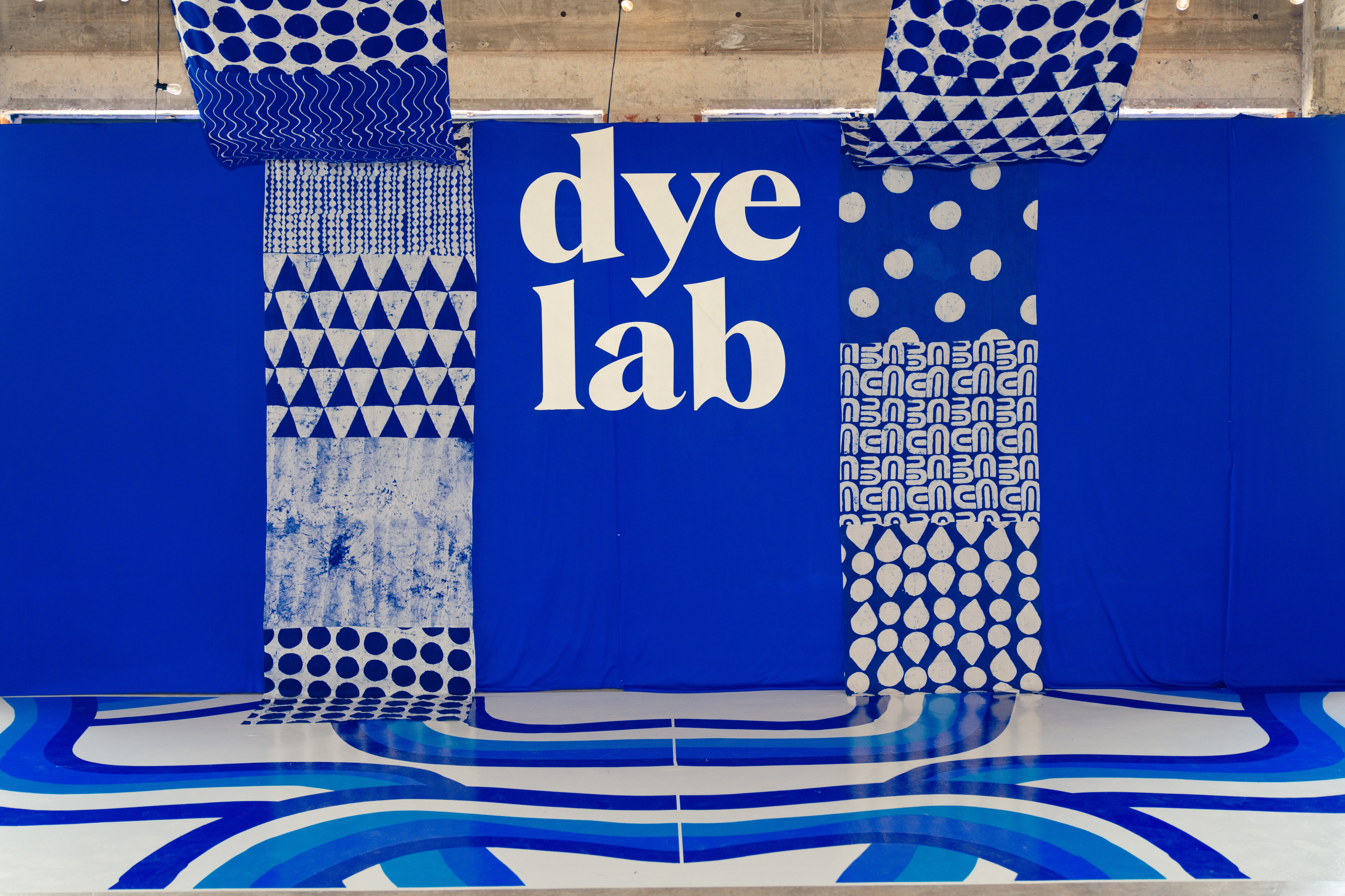 Blue wall with white text 'dye lab' and patterned fabric samples hanging, with a wavy blue and white design on the floor.