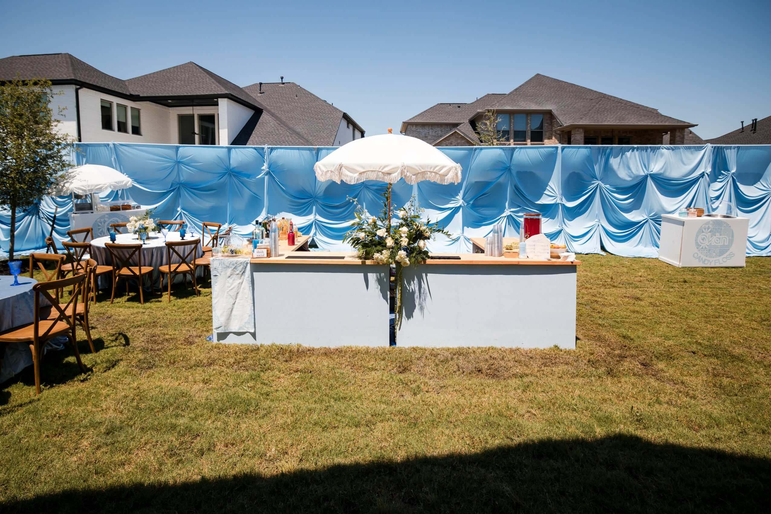 Outdoor party setup with tables, chairs, umbrellas, and a decorated backdrop on a grassy area.