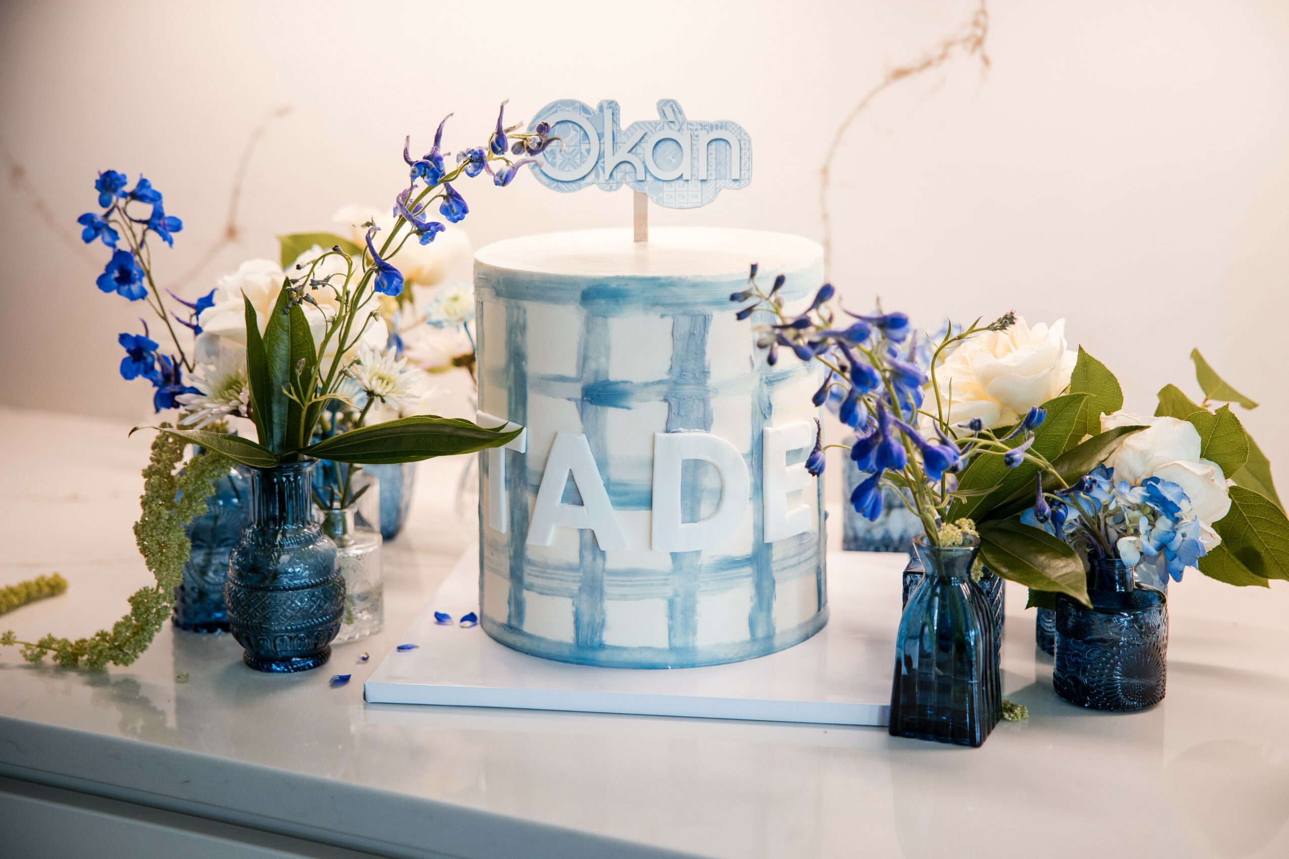 A birthday cake with blue and white design, surrounded by blue and white flowers in vases, on a table.