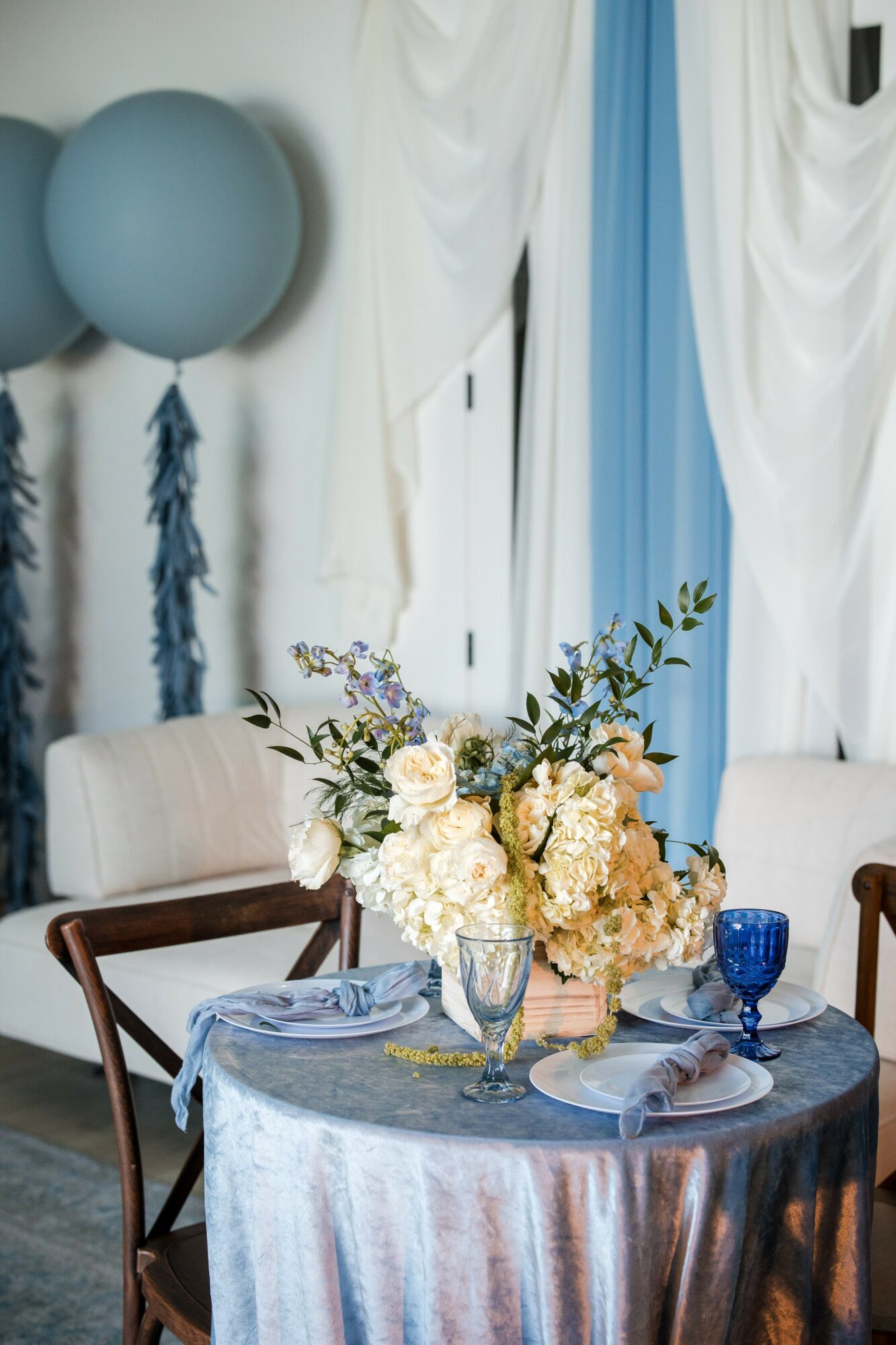 Table with floral centerpiece, set with plates, glasses, and blue and white decorations, in a decorated room with curtains and balloons.