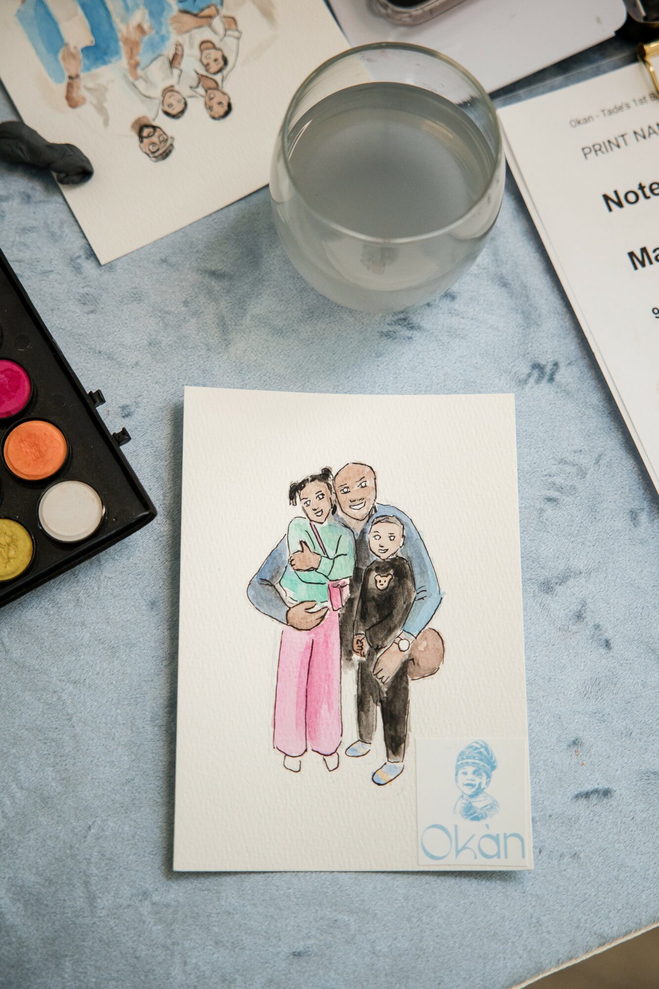 Hand-drawn illustration of three people, one holding a book, on a white card on a table with a glass of water and papers