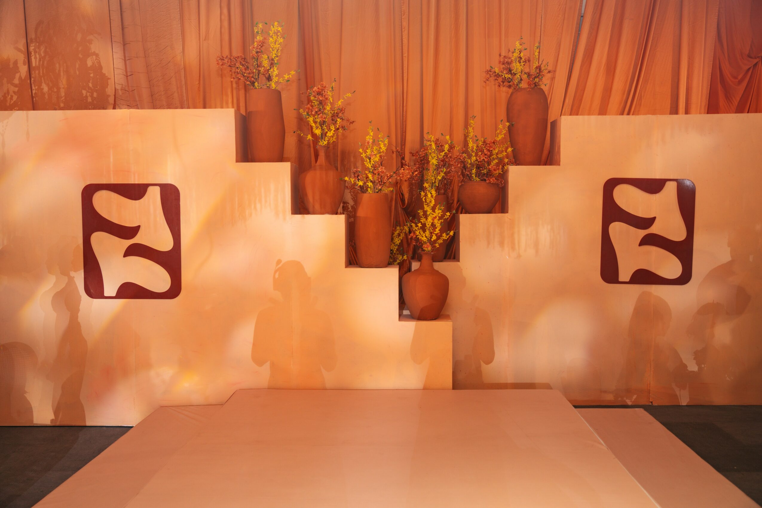 Stage with decorative vases and flowers, backdrop with warm lighting, and abstract designs on panels.