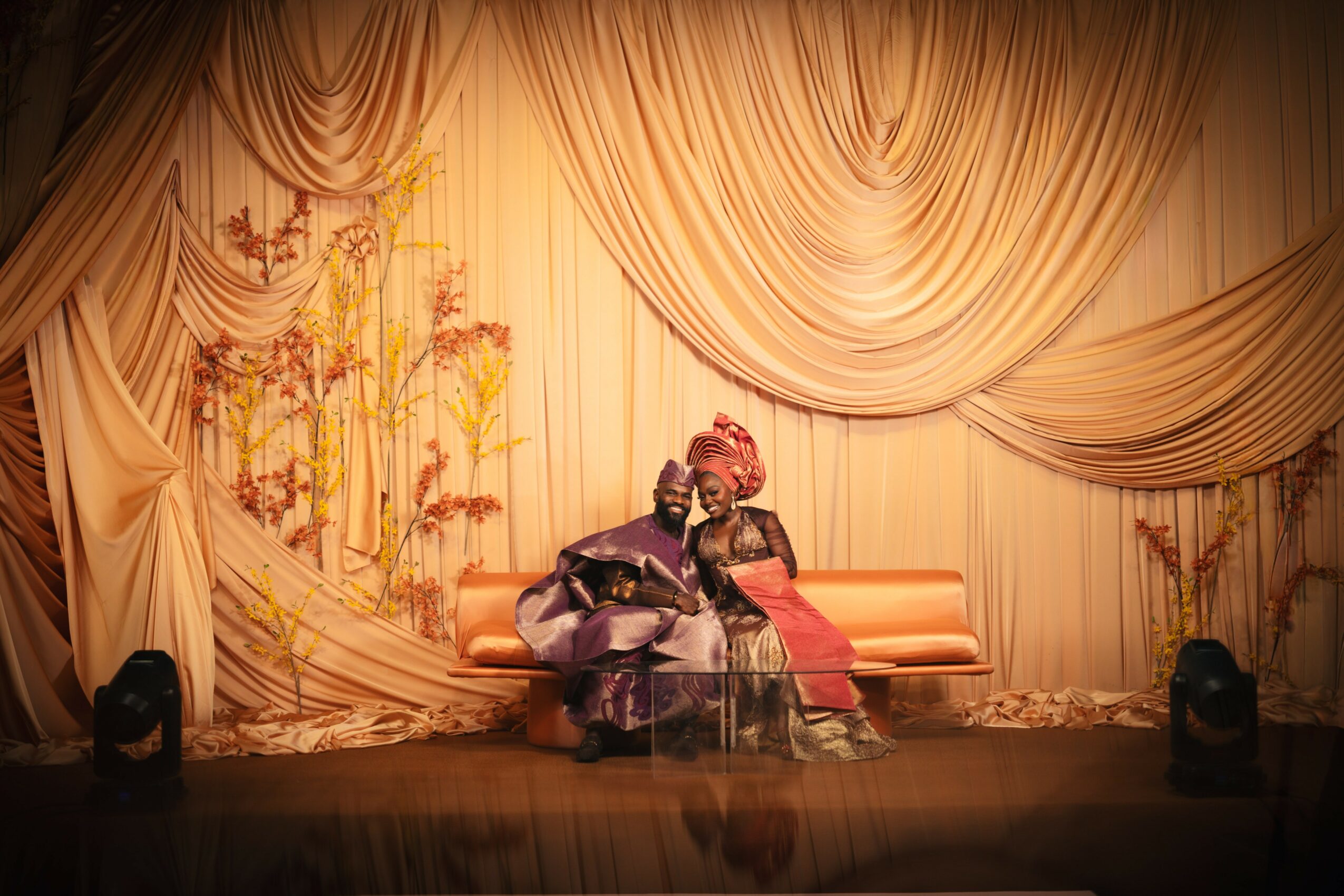 Two performers in traditional costumes sit on a stage with a decorated curtain backdrop, floral designs, and stage lights.