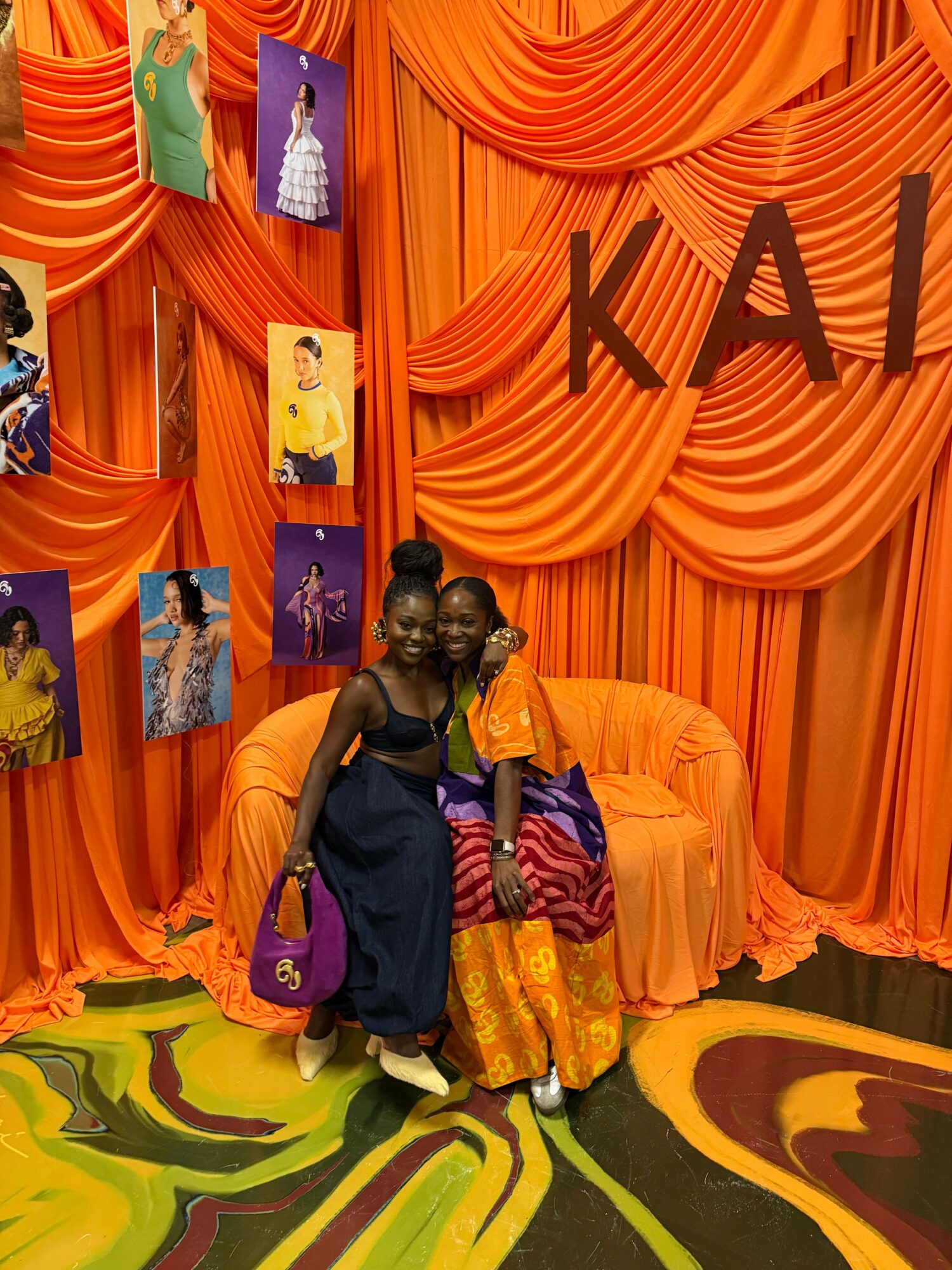 Three people stand in front of a colorful backdrop with orange, yellow, and purple patterns, holding bags and wearing casual clothing.