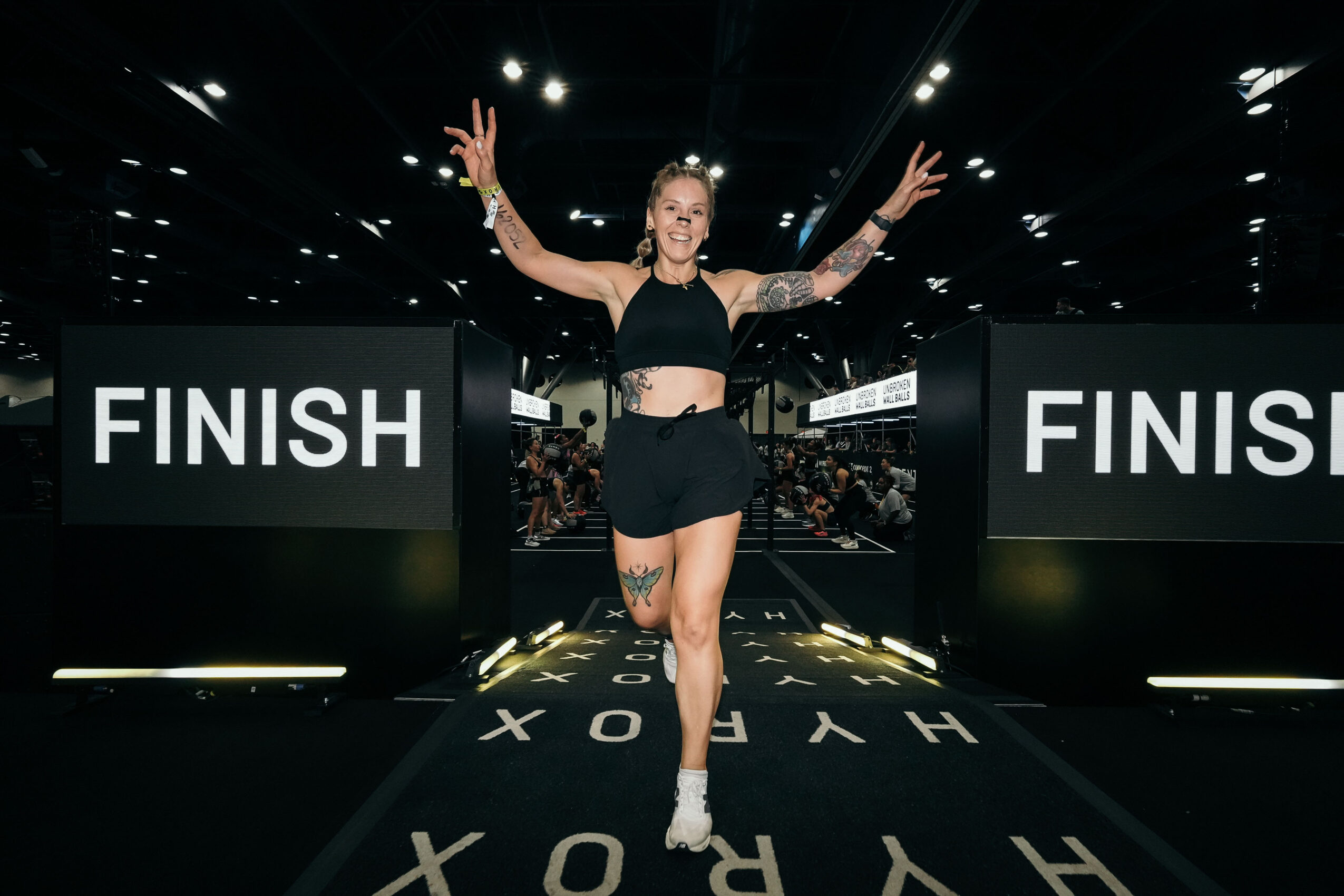 A woman crosses the finish line with arms raised in a large indoor event, smiling, with a crowd in the background.
