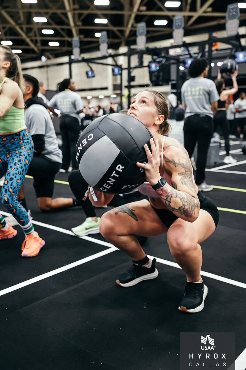 Woman with tattoos holding a medicine ball in a gym, surrounded by people and workout equipment, in a squat position.