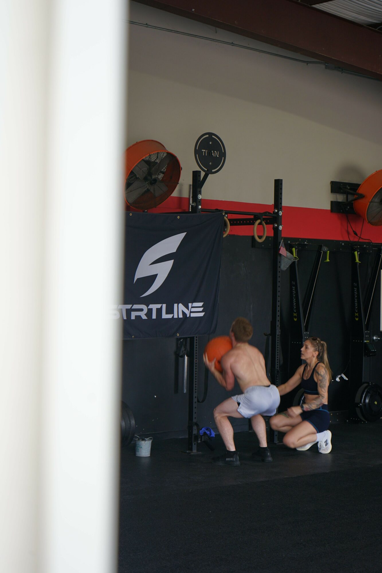 Person with red hair sitting on the floor in a gym, lifting a barbell with weights, surrounded by gym equipment.