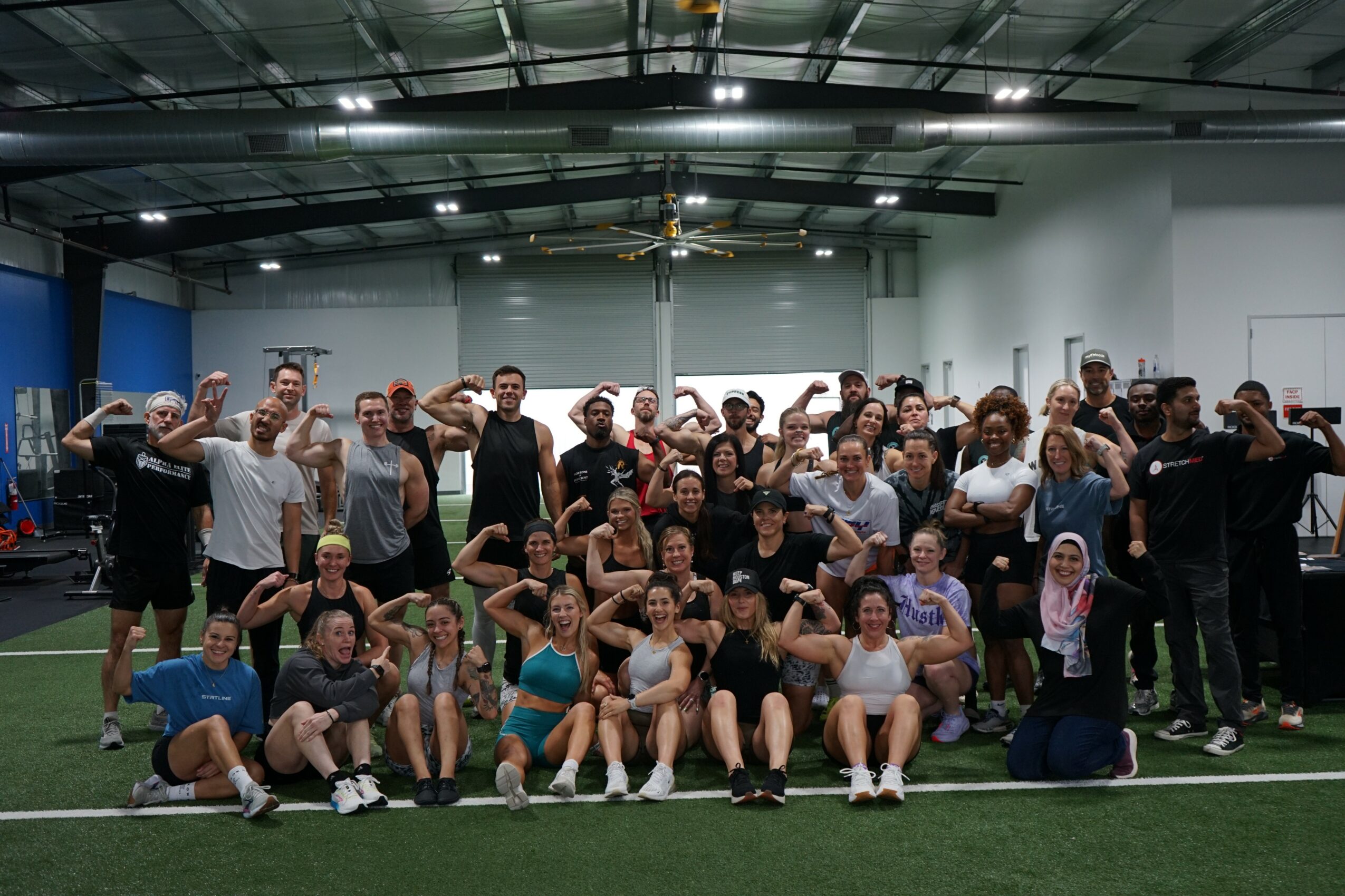 Group of people posing together indoors on a sports field, some sitting and some standing, smiling and making gestures.