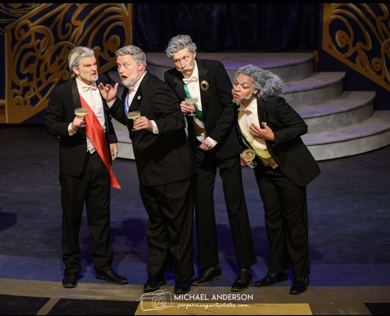 Four people in tuxedos stand on stage, holding drinks and singing, with a staircase in the background.