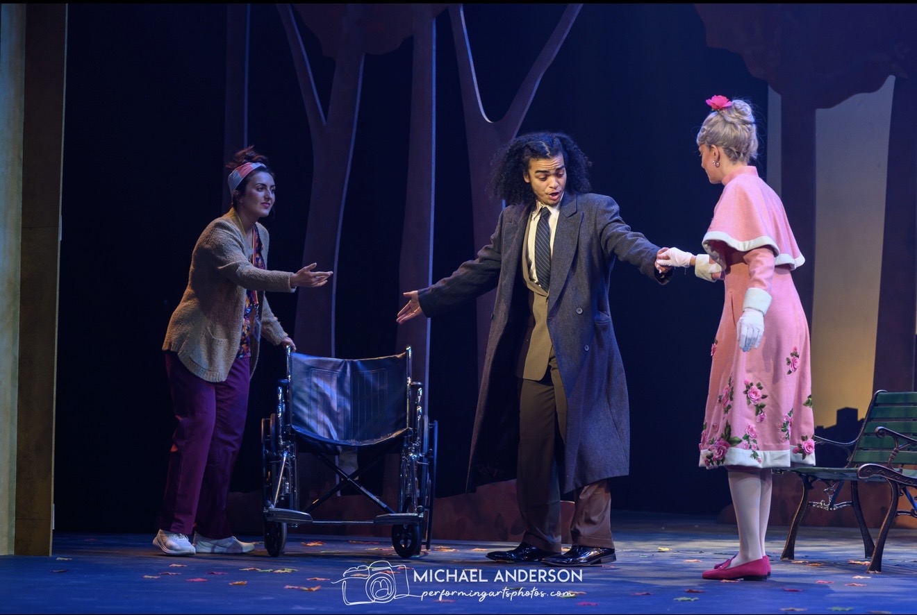 Three women on stage, one holding hands with another, with a chair and a backdrop of trees, performing a scene.