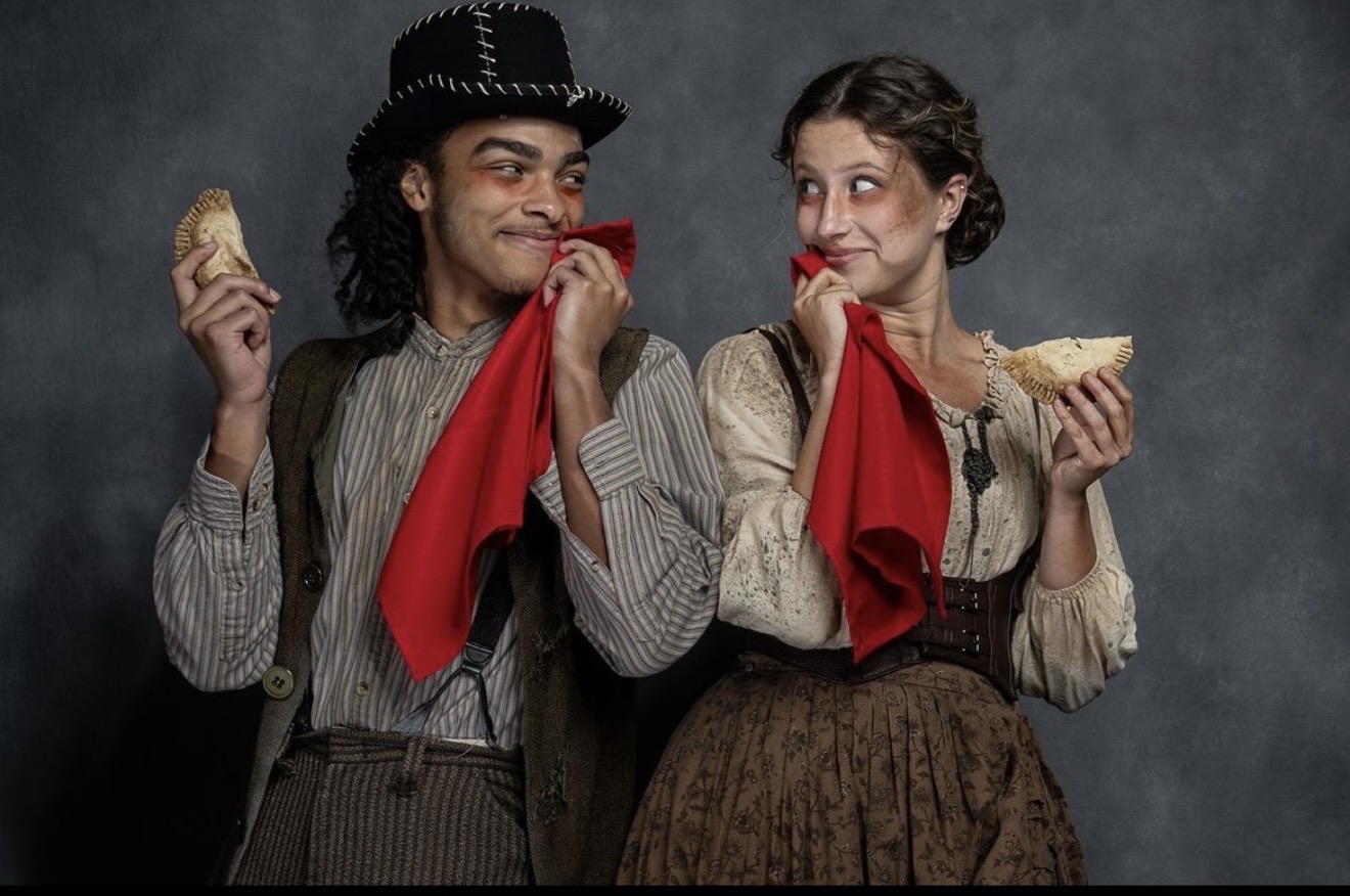 Two people dressed in vintage clothing holding bread and red cloths, smiling at each other, against a dark background.